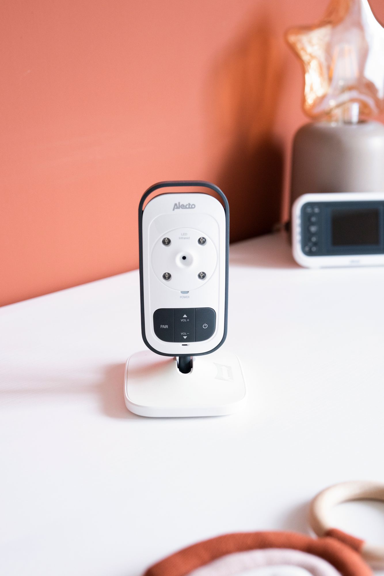 Niania elektroniczna na białym stole. Monitor jest biały z czarnym przodem. Tło to ściana i dekoracje.