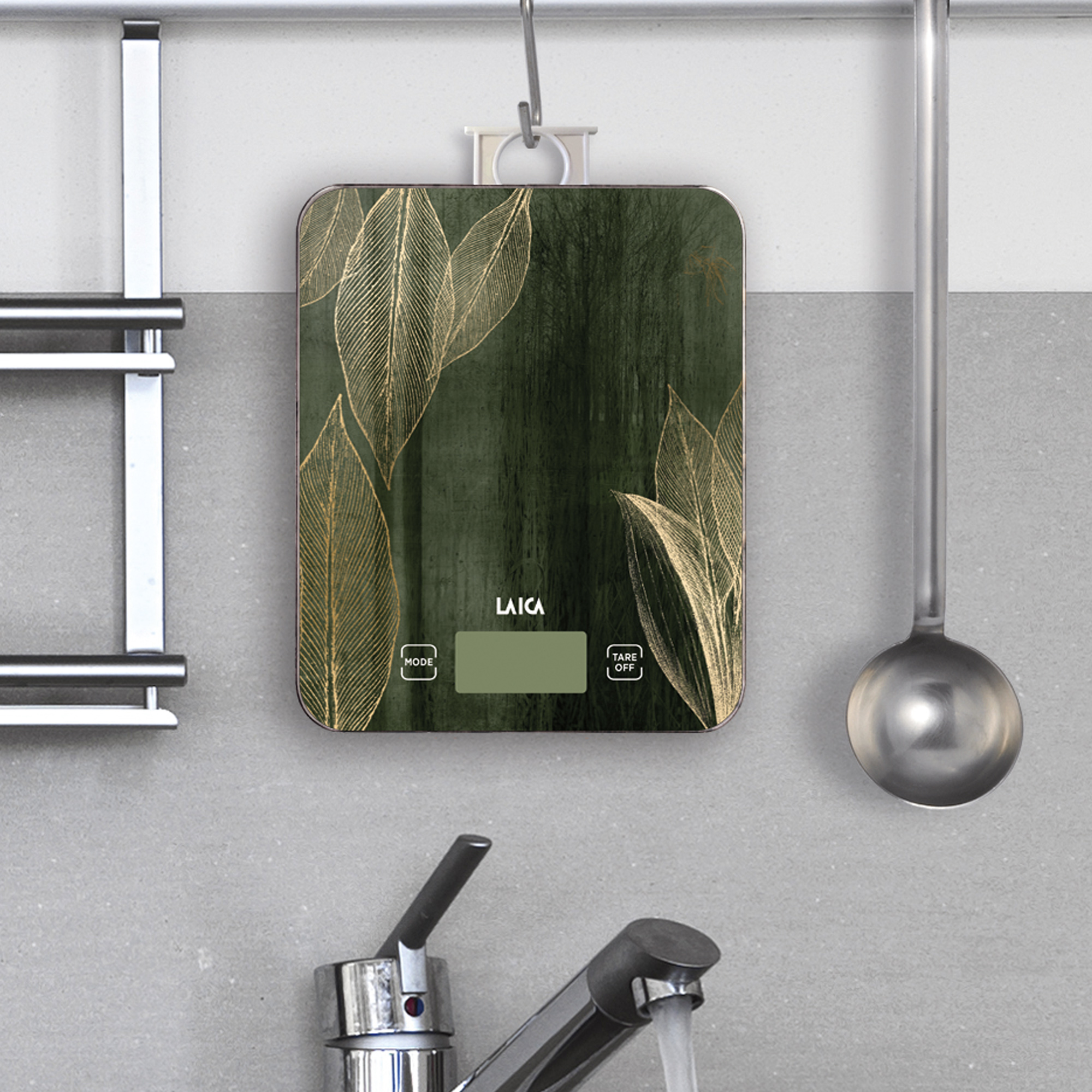 Una báscula de cocina digital verde con hojas doradas, colgada en una pared de azulejos grises.