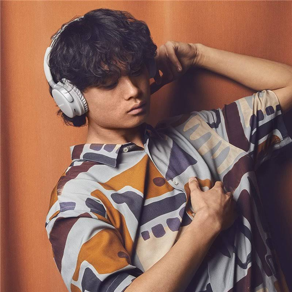 Un joven con auriculares y una camisa estampada se apoya en una pared naranja.