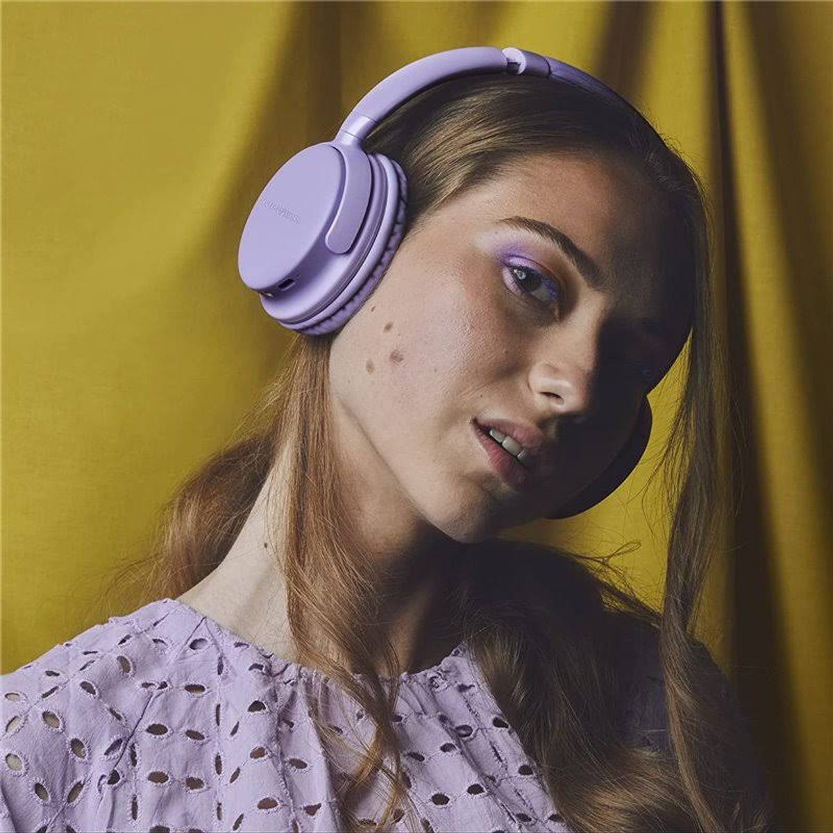 Una mujer con auriculares morados. Ella está mirando a la cámara. Fondo amarillo.