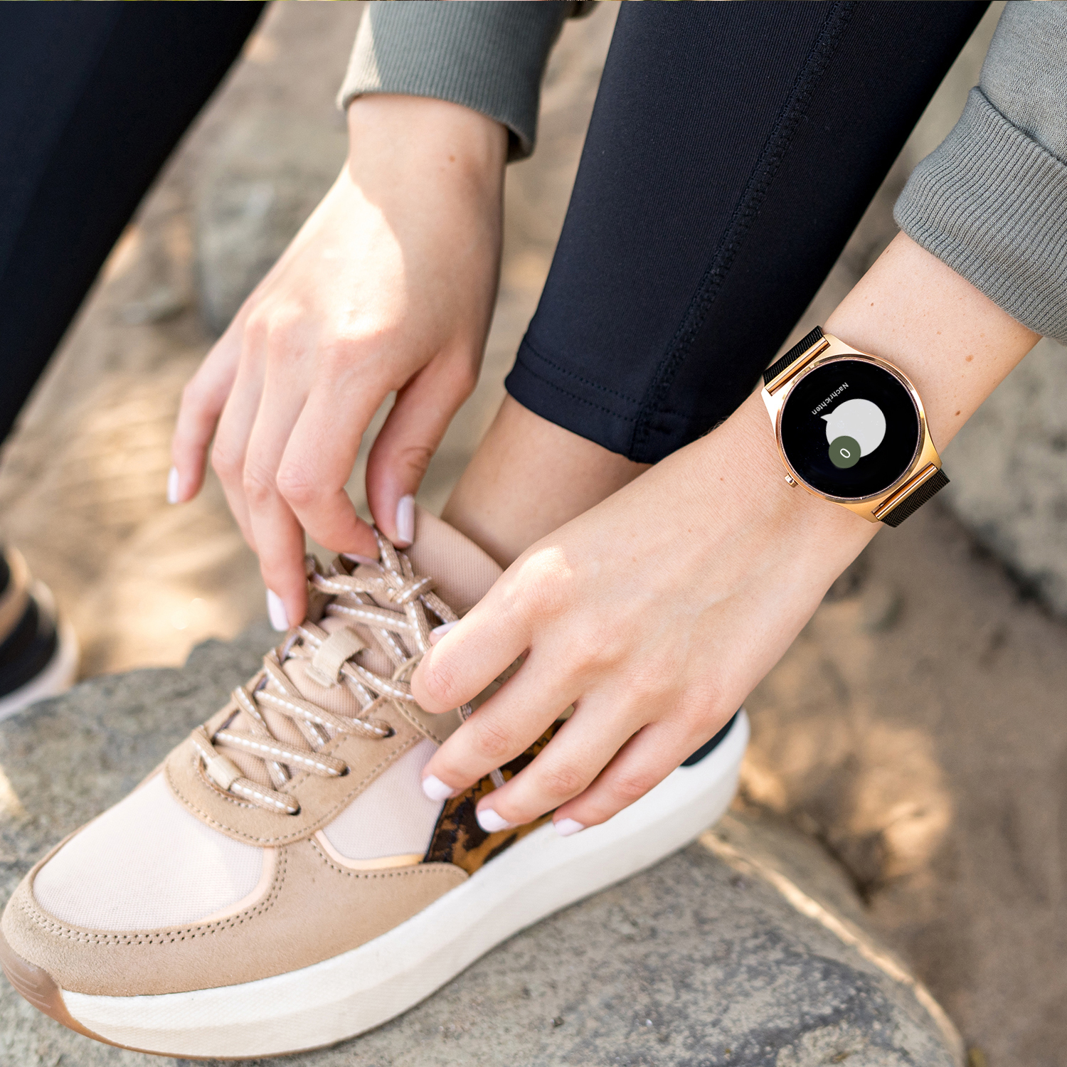Eine Frau bindet ihren Schuh, trägt eine Smartwatch. Die Uhr ist golden. Beige Turnschuhe.