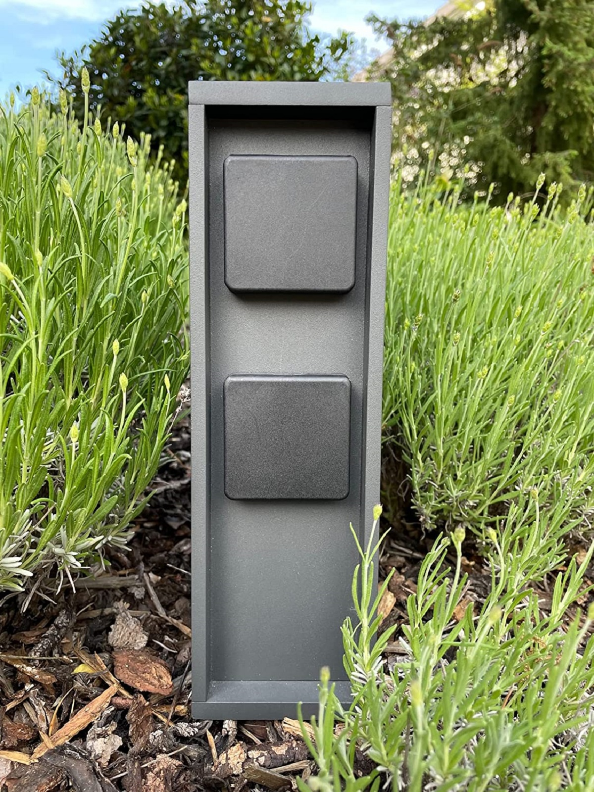 Dunkelgraue Steckdosenbox mit zwei schwarzen Steckdosen, umgeben von grünen Pflanzen. Außenbereich.
