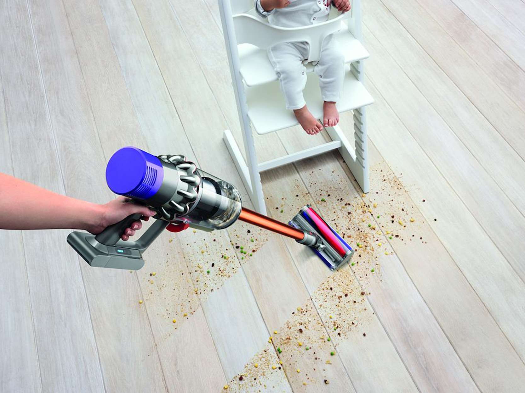 Eine Hand, die einen Staubsauger über einem Holzboden hält, mit einem Baby, das in einem Hochstuhl sitzt.