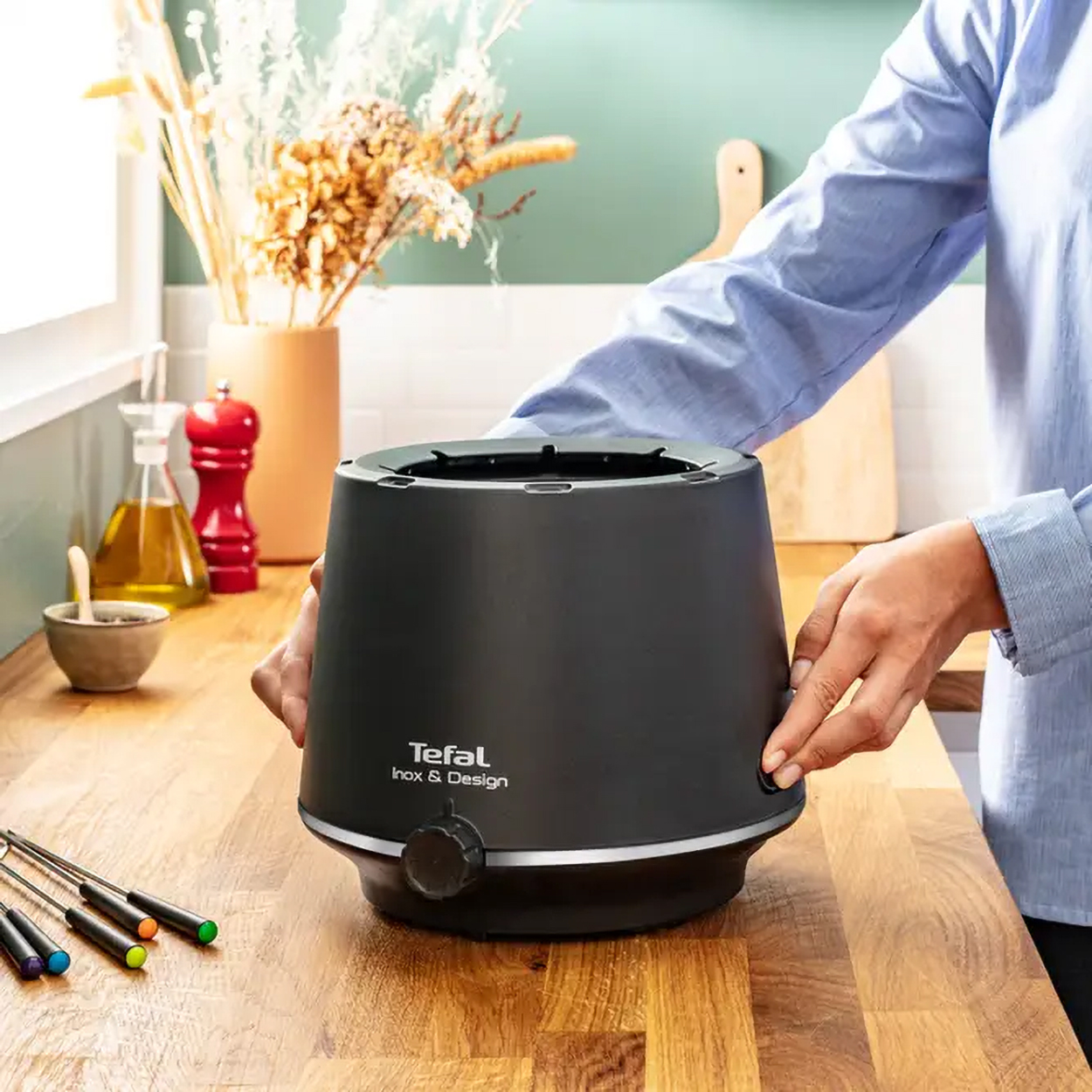 Czarny garnek do fondue ze srebrną gałką na drewnianej powierzchni. Kwiaty i inne przedmioty w tle.