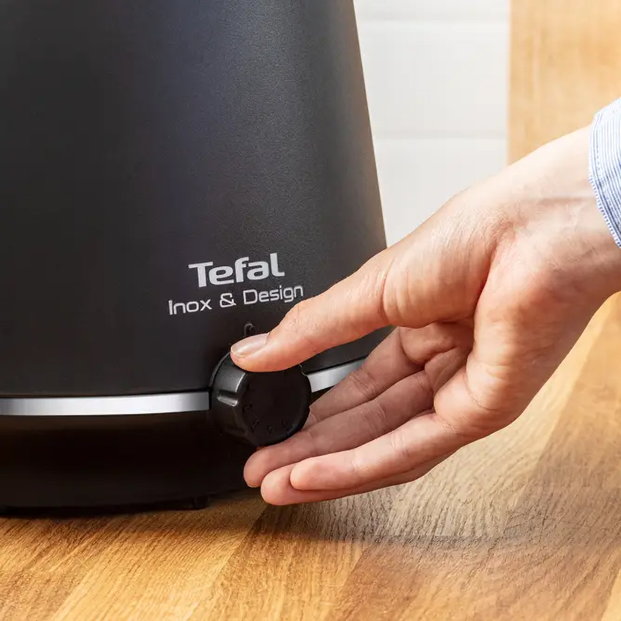 Ręka kręci czarnym pokrętłem na czarnym urządzeniu kuchennym z logo 'Tefal'. Drewniana powierzchnia.