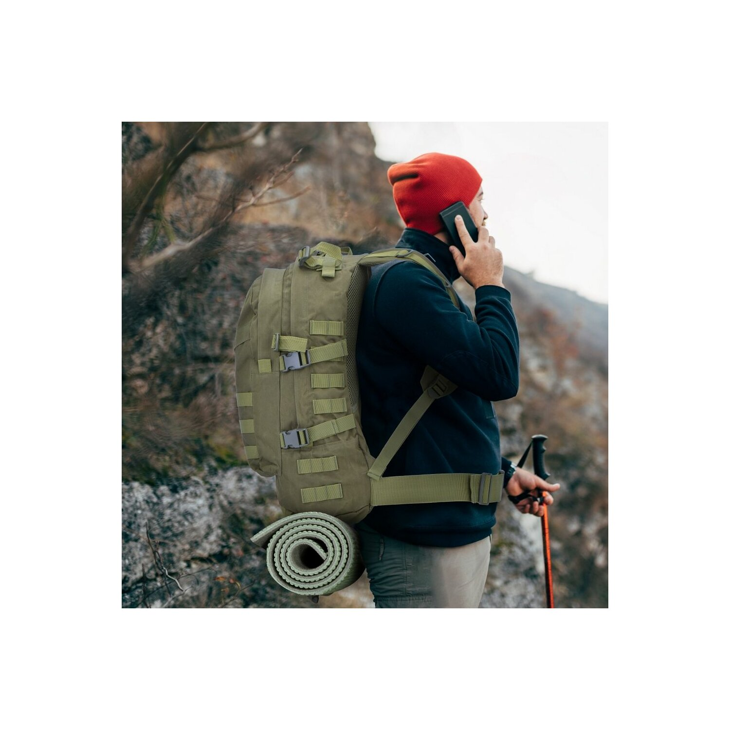 Ein Mann mit roter Mütze und Rucksack auf felsigem Hügel, telefoniert, mit Matte.