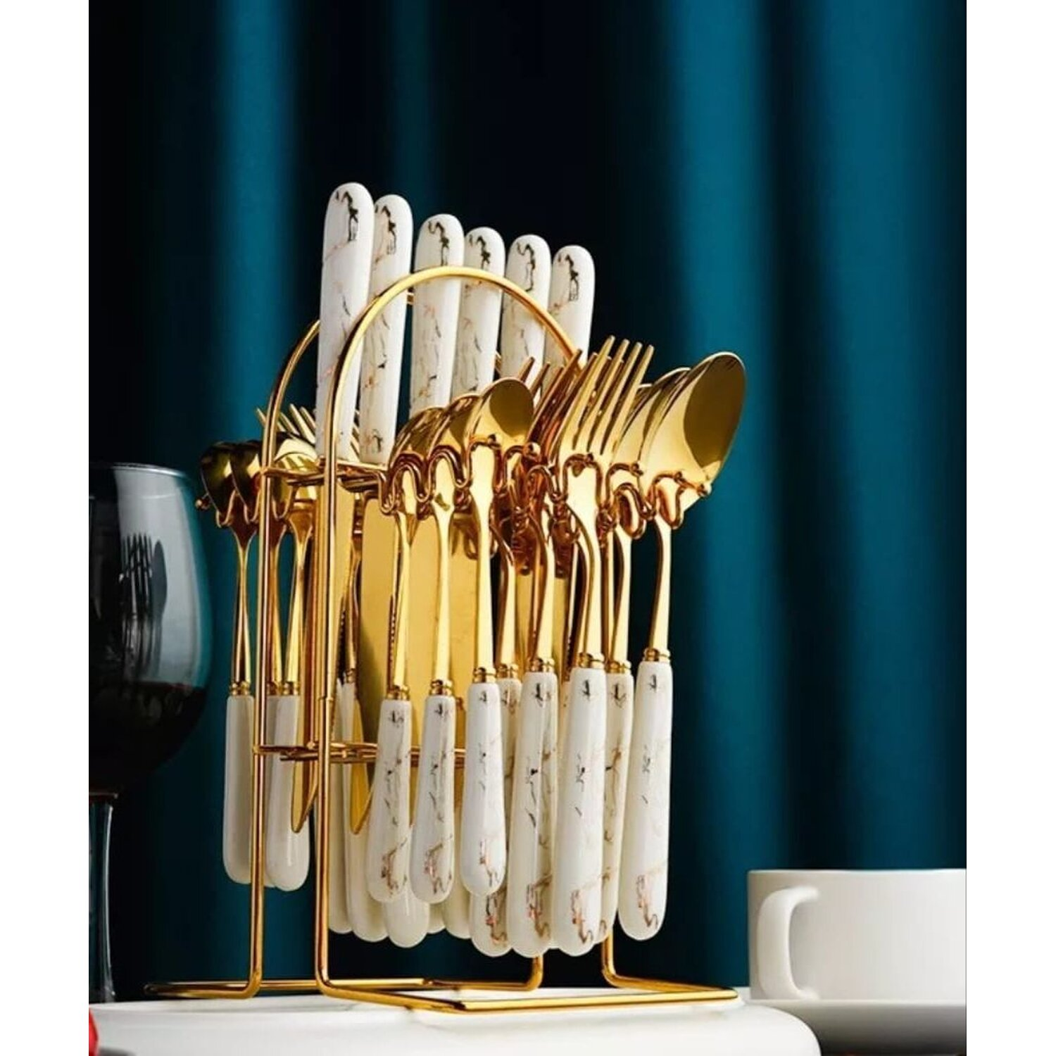 Goldenes Besteck-Set mit weißen Marmorgriffen in einem Goldhalter. Blauer Vorhang im Hintergrund. Glas und weiße Tasse an der Seite.