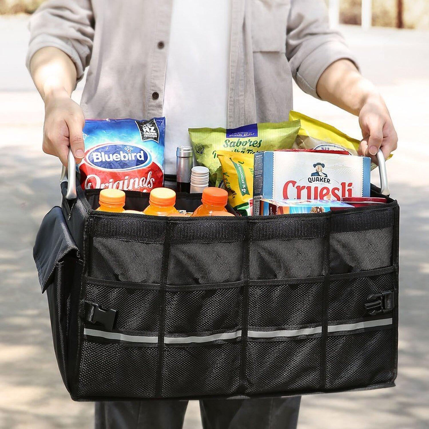 Person hält einen schwarzen Kofferraum-Organizer voller Lebensmittel: Saft, Snacks und Kartons.