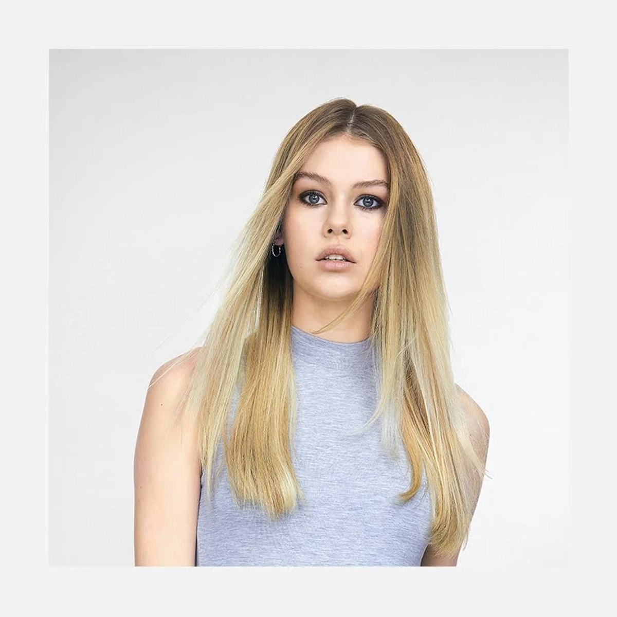 Kobieta z długimi, blond włosami, patrząca w kamerę, ubrana w szary top.