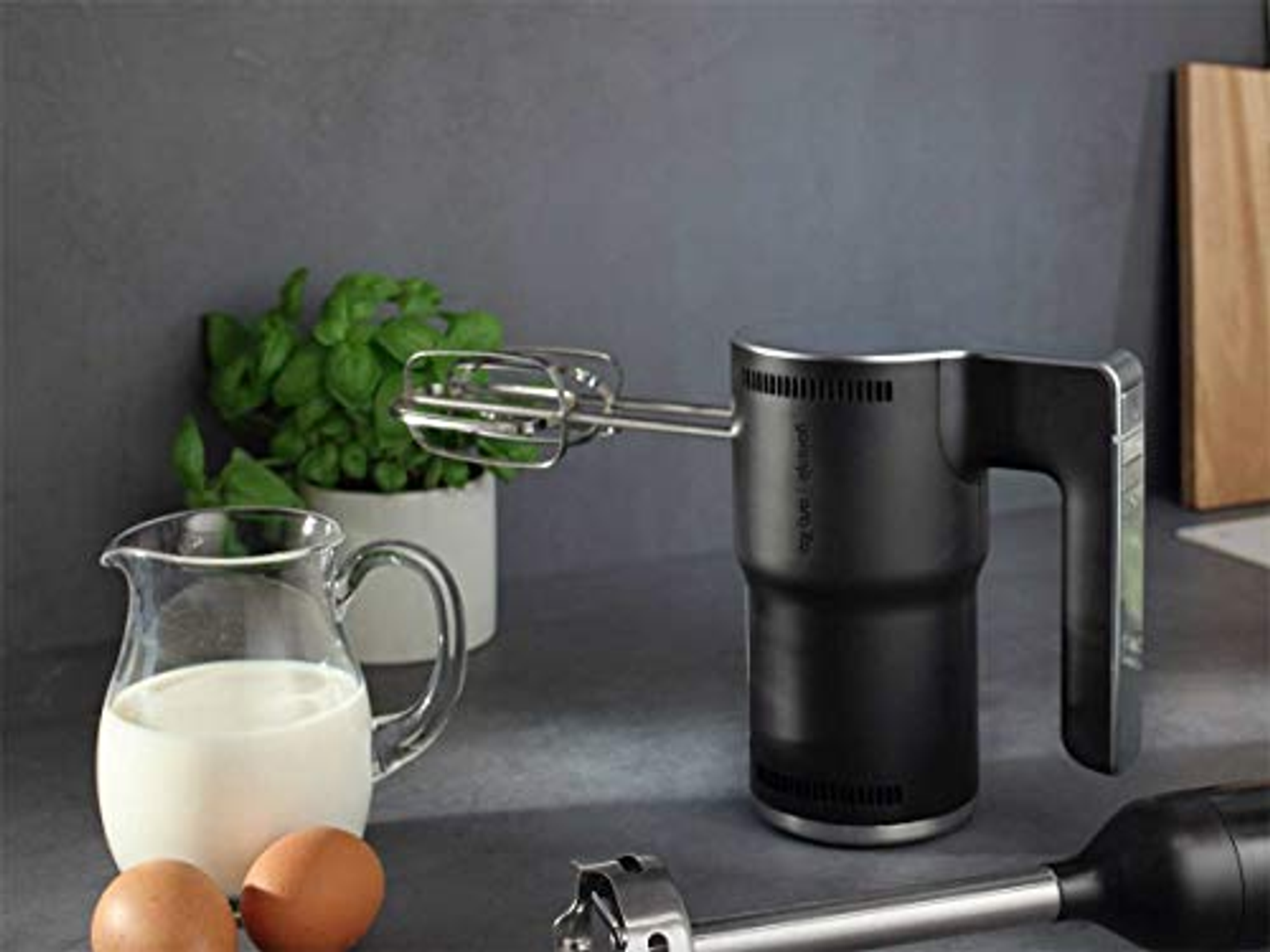 Schwarzer Handmixer mit Aufsätzen, Eiern, Milch und Pflanze auf grauer Oberfläche.
