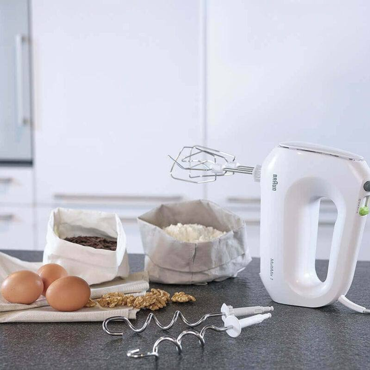 Weißer Handmixer mit Zubehör neben Mehl, Eiern und Nüssen auf einem Tisch.