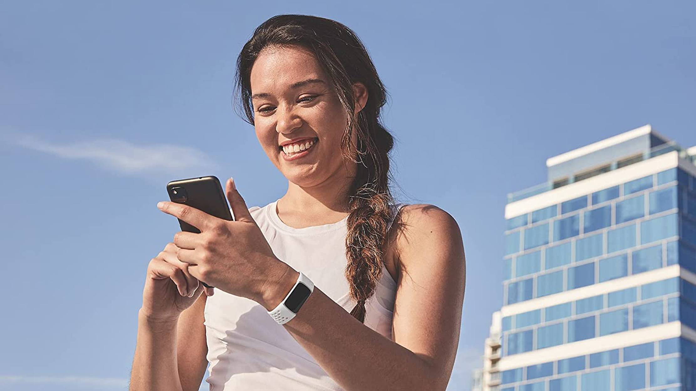 Eine Frau lächelt, während sie ein Smartphone benutzt. Sie trägt eine Uhr, mit einem Gebäude hinter ihr.