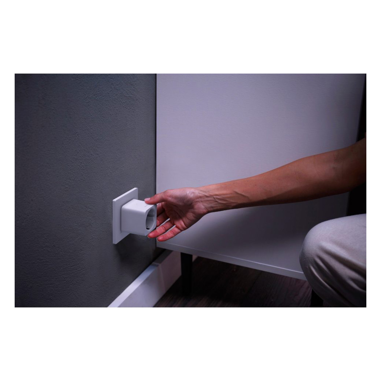 Hand inserting a white smart plug into an outlet on a gray wall.