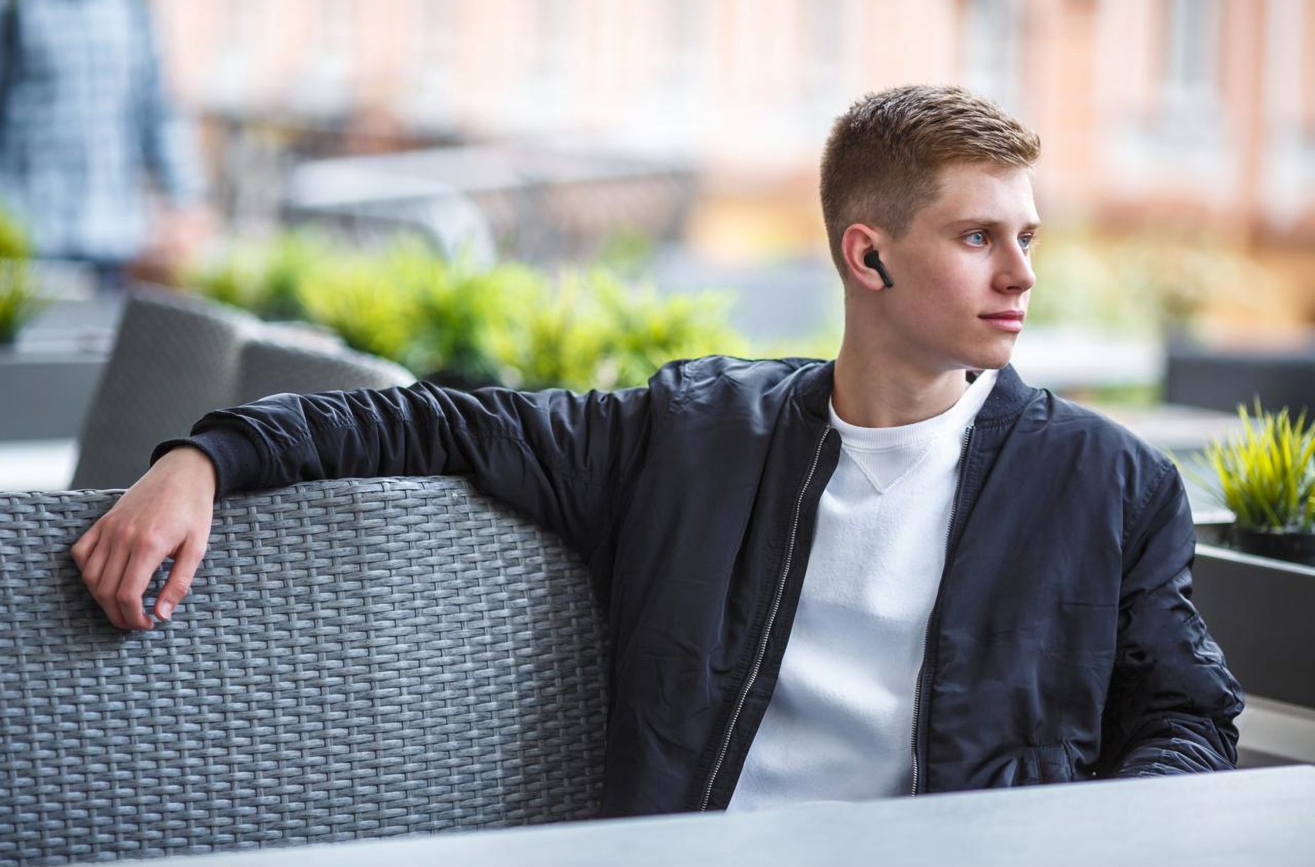 Młody mężczyzna z czarnymi słuchawkami siedzi na zewnątrz. Ma na sobie czarną kurtkę i patrzy w bok.