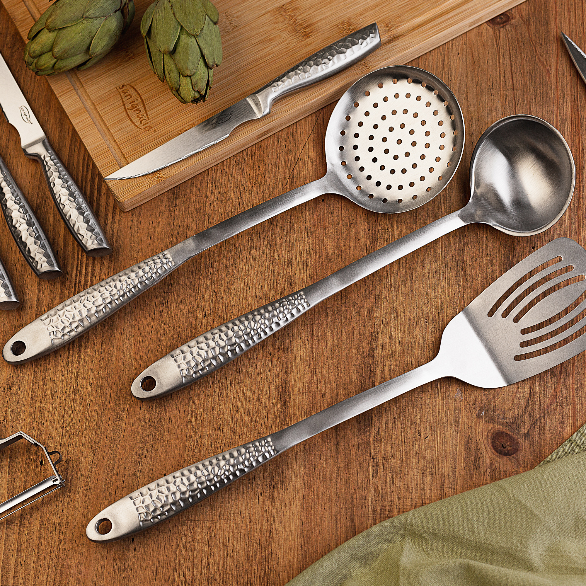 Una mesa de madera con utensilios de cocina: una cuchara ranurada, un cucharón, una espátula y cuchillos, con alcachofas.