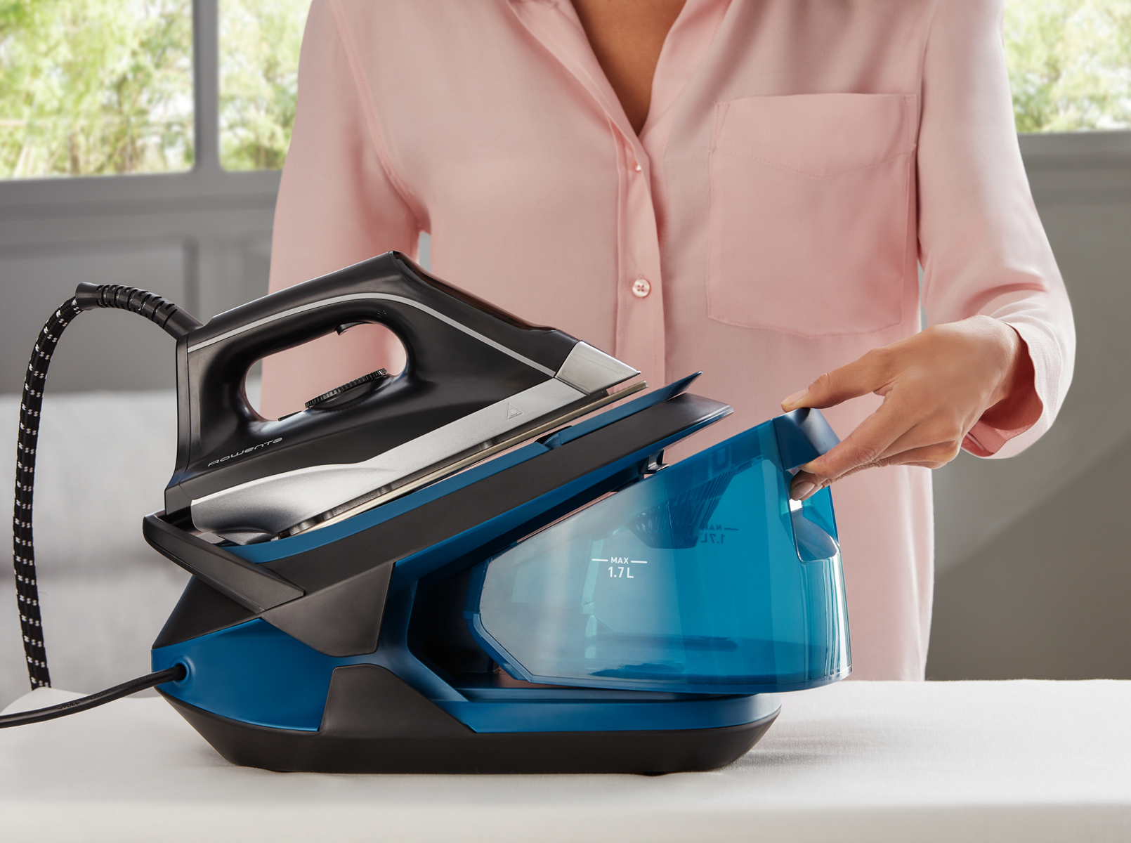 Frau befüllt blauen Wassertank eines schwarz-blauen Rowenta-Bügeleisens auf weißer Fläche.