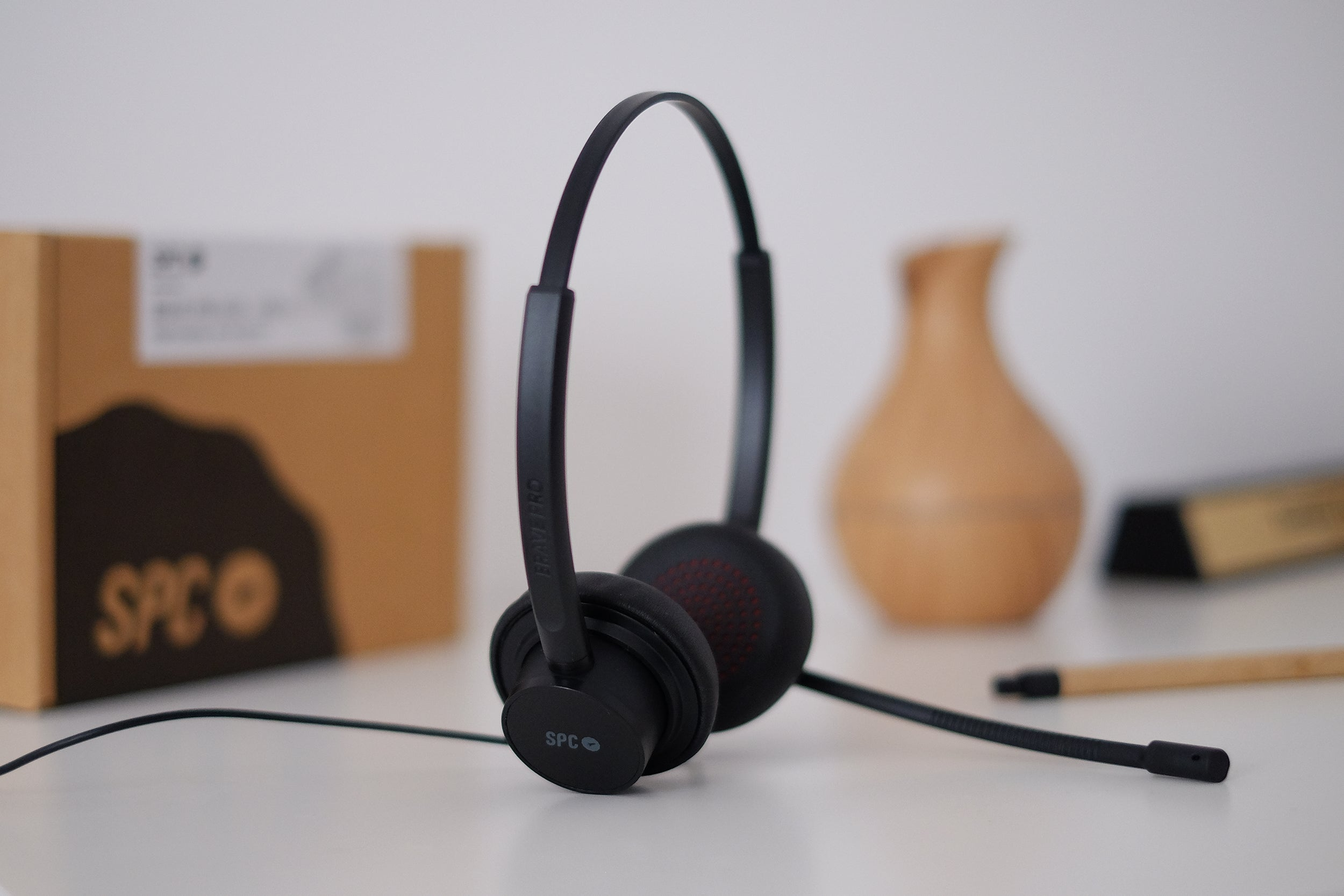 Auriculares negros sobre una mesa blanca, con una caja marrón y un jarrón al fondo.
