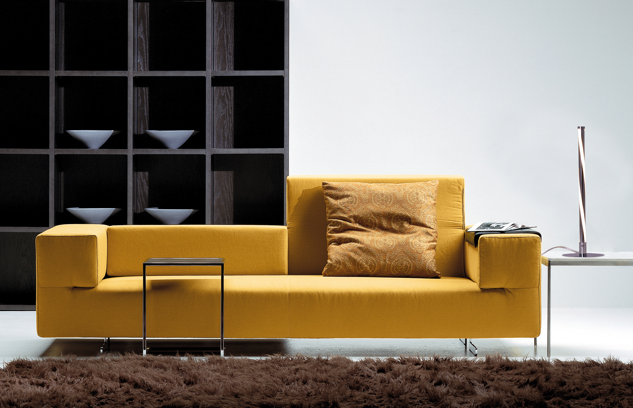 Gelbes Sofa mit Tisch und Lampe. Dunkles Regal im Hintergrund.