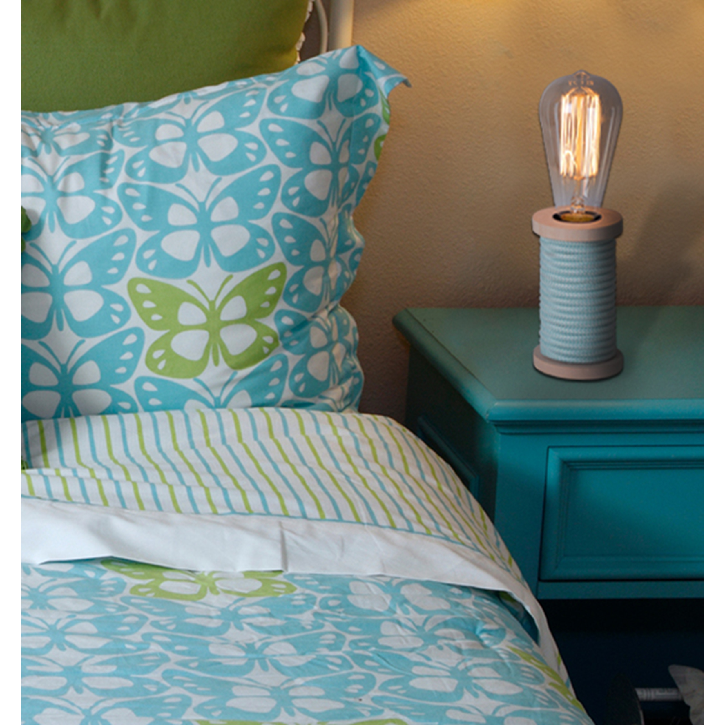 Bett mit blau-grün gemusterter Bettdecke, türkisfarbenem Nachttisch und Lampe.
