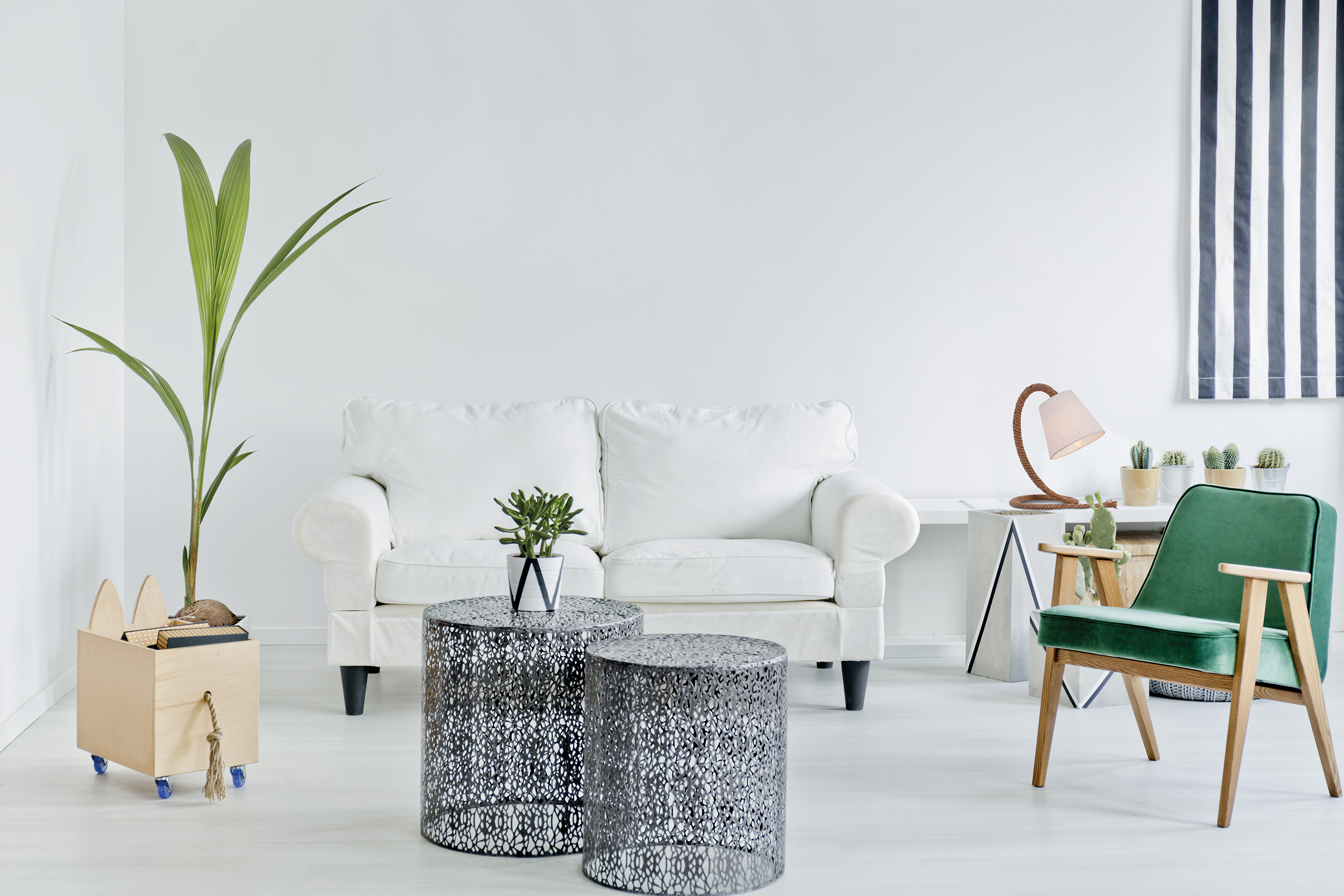 Weißes Sofa, zwei Metalltische, grüner Stuhl, Pflanze in Holzkiste, weiße Wand im Raum.