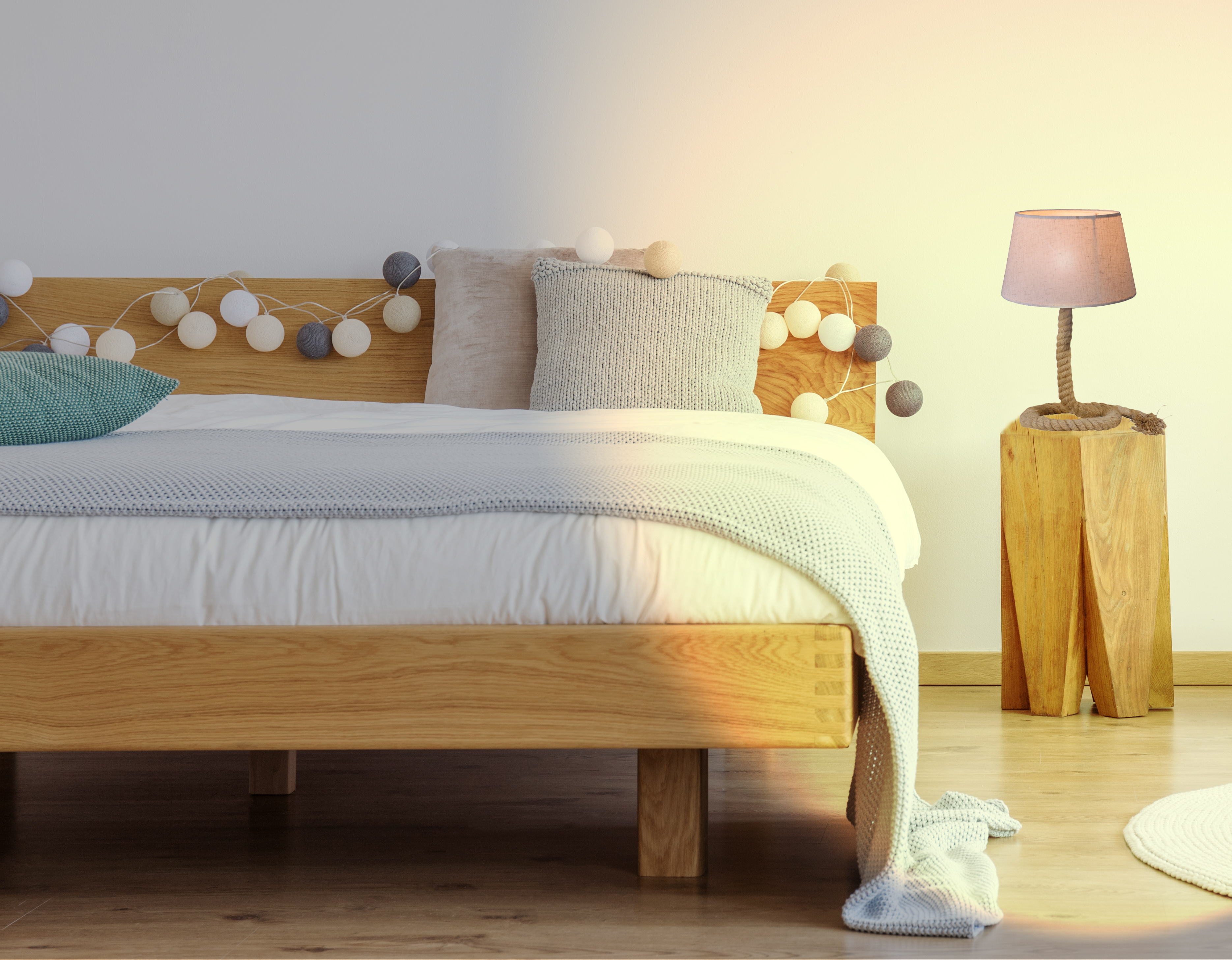 Ein Bett mit Kissen, einer Decke und dekorativen Lichtern. Eine kleine Lampe steht auf einem Nachttisch aus Holz.