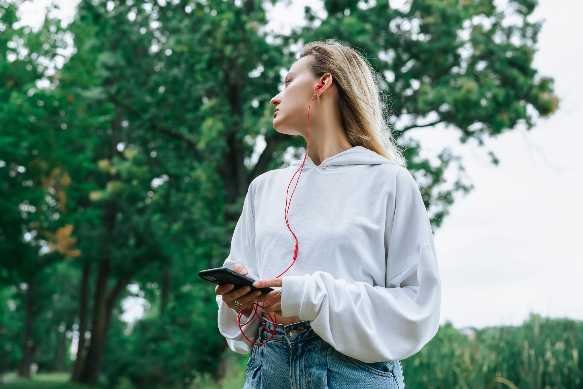 Blonde Frau mit Kopfhörern, hält ein Telefon, schaut weg. Bäume im Hintergrund.