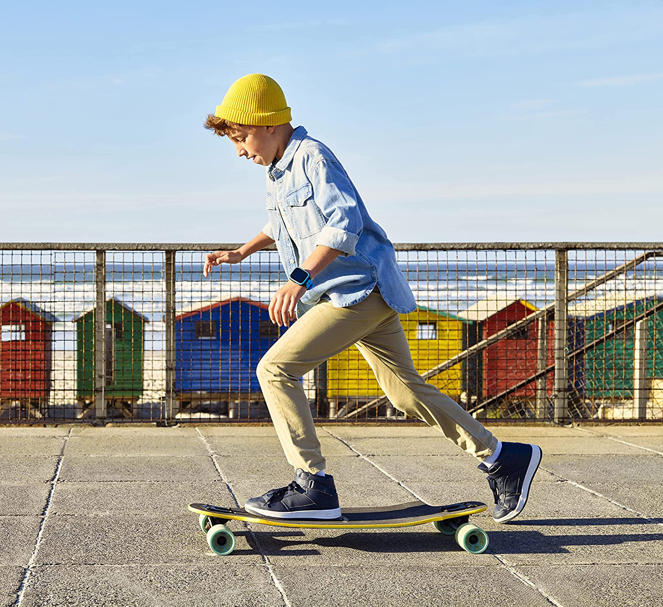 Junge mit gelbem Hut fährt auf einem gelben Skateboard in der Nähe farbiger Strandhütten.