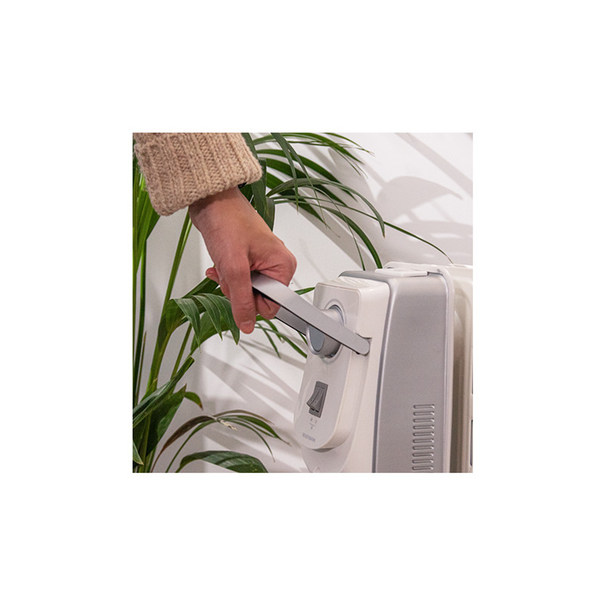 Osoba trzyma grzejnik. Zielona roślina w tle. Biała ściana.