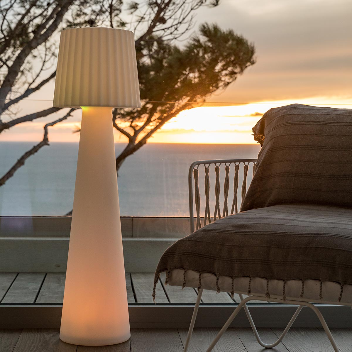 Una lampada bianca alta con paralume, su un ponte di legno con vista sul tramonto.