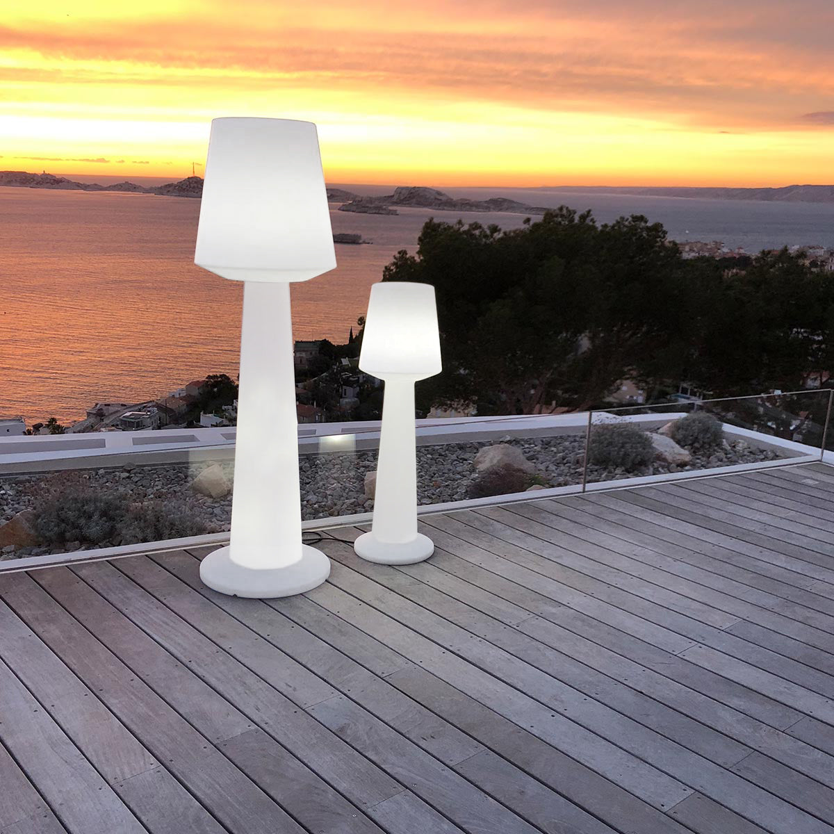 Zwei weiße Außenlampen auf Holzterrasse. Sonnenuntergangshimmel im Hintergrund, mit Wasser und Land.
