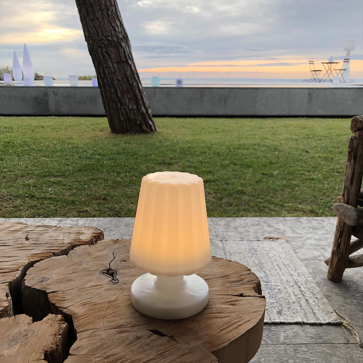 Eine weiße, kleine Tischlampe leuchtet auf einem Holztisch, mit malerischem Hintergrund von Meer und Himmel.