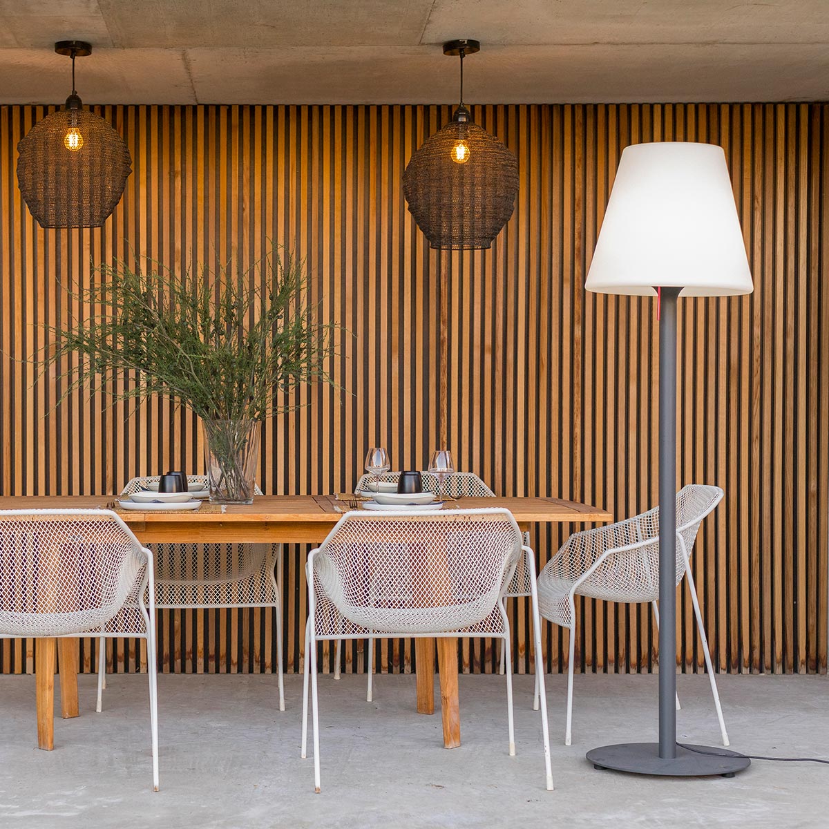 Sala da pranzo all'aperto con due lampade a sospensione. Tavolo apparecchiato, sedie e lampada da terra.