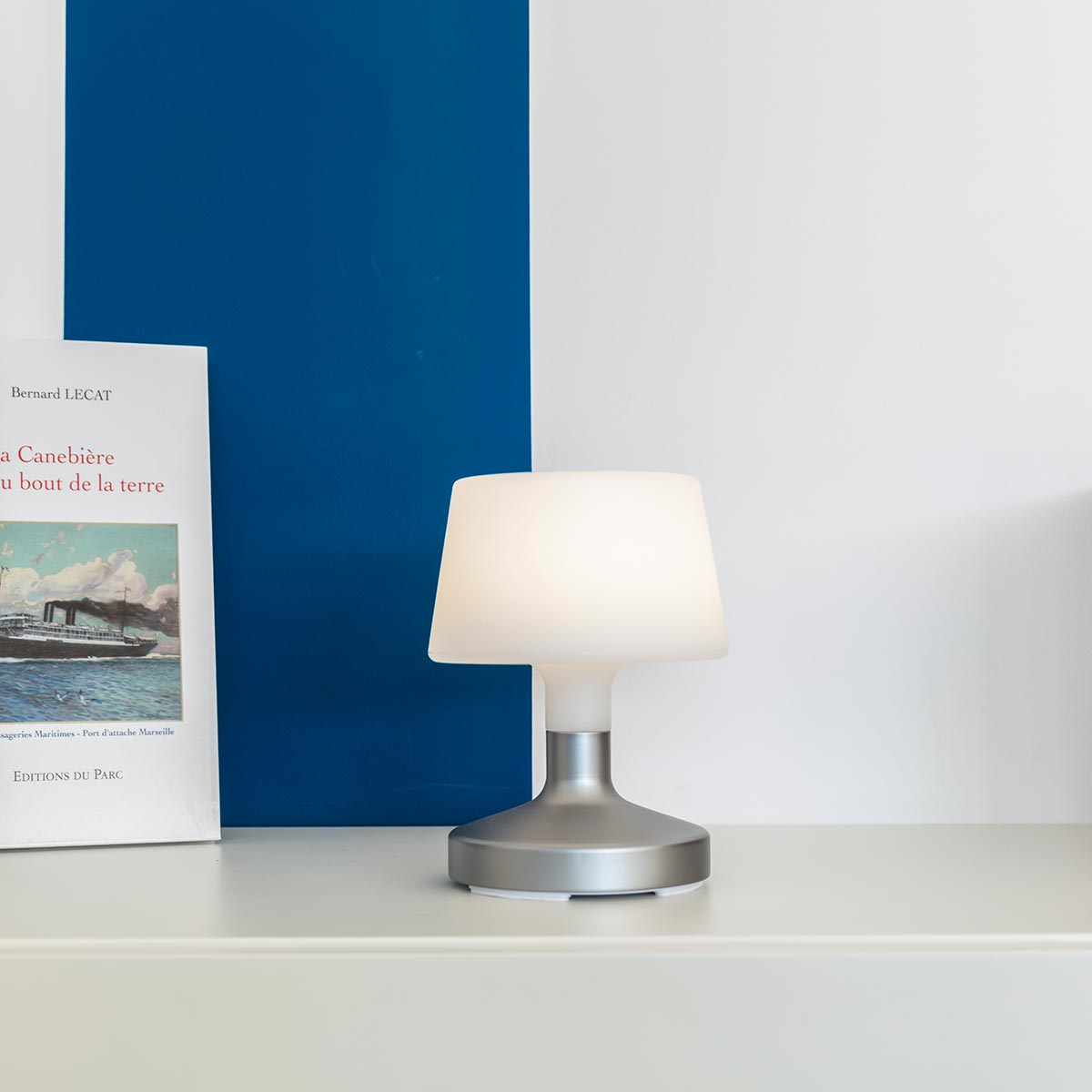 Weiße und silberne Tischlampe mit weißem Lampenschirm auf weißer Oberfläche. Blaue Wand dahinter.