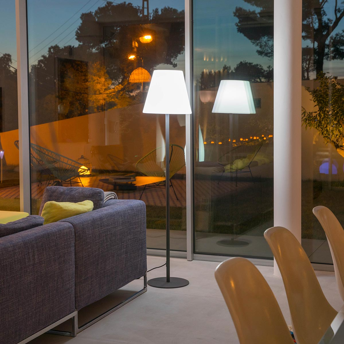 Ein Wohnzimmer mit Stehlampe. Ein graues Sofa und Stühle sind sichtbar. Nachtansicht.