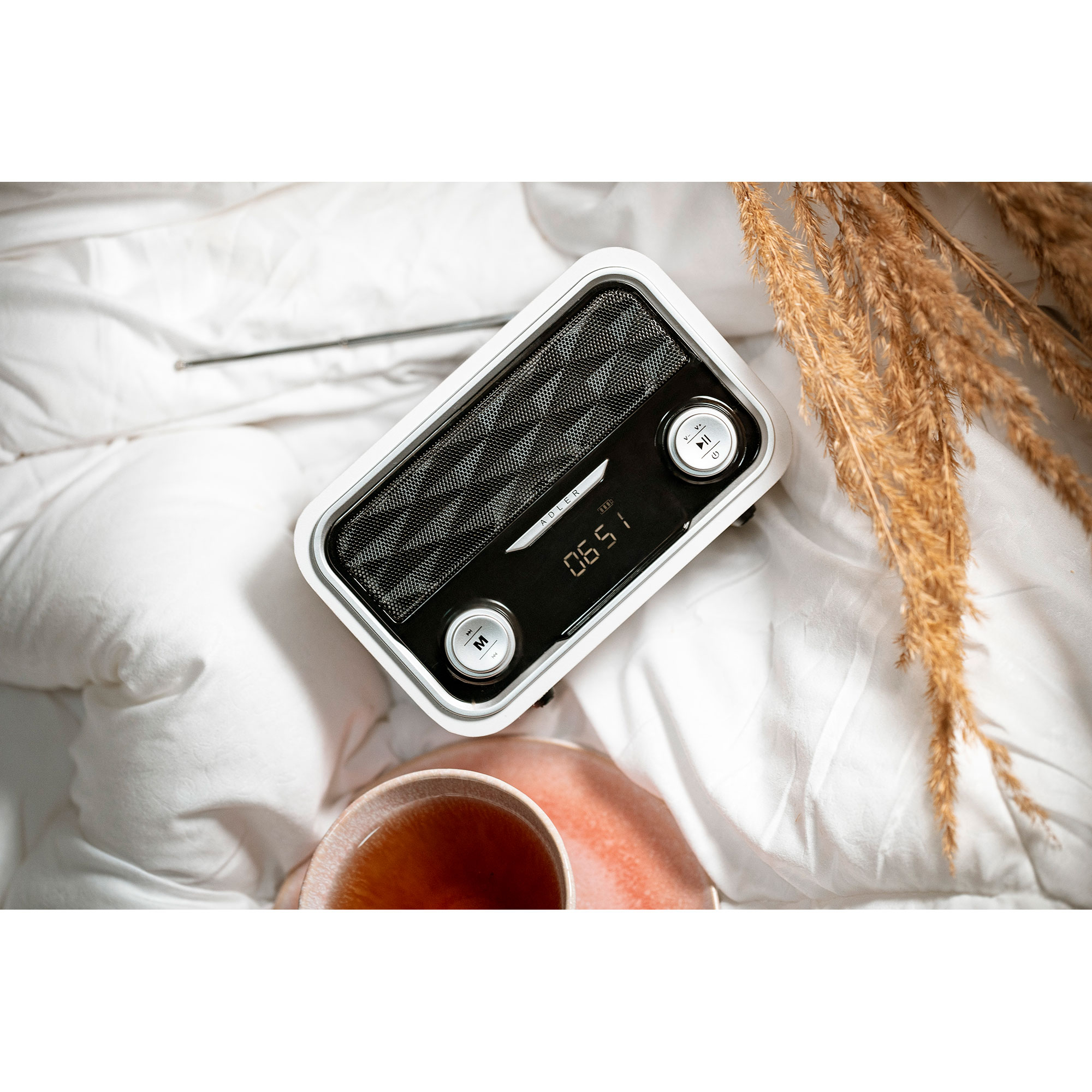 Radio bianca con display digitale, una tazza di tè e piante essiccate su un letto bianco.