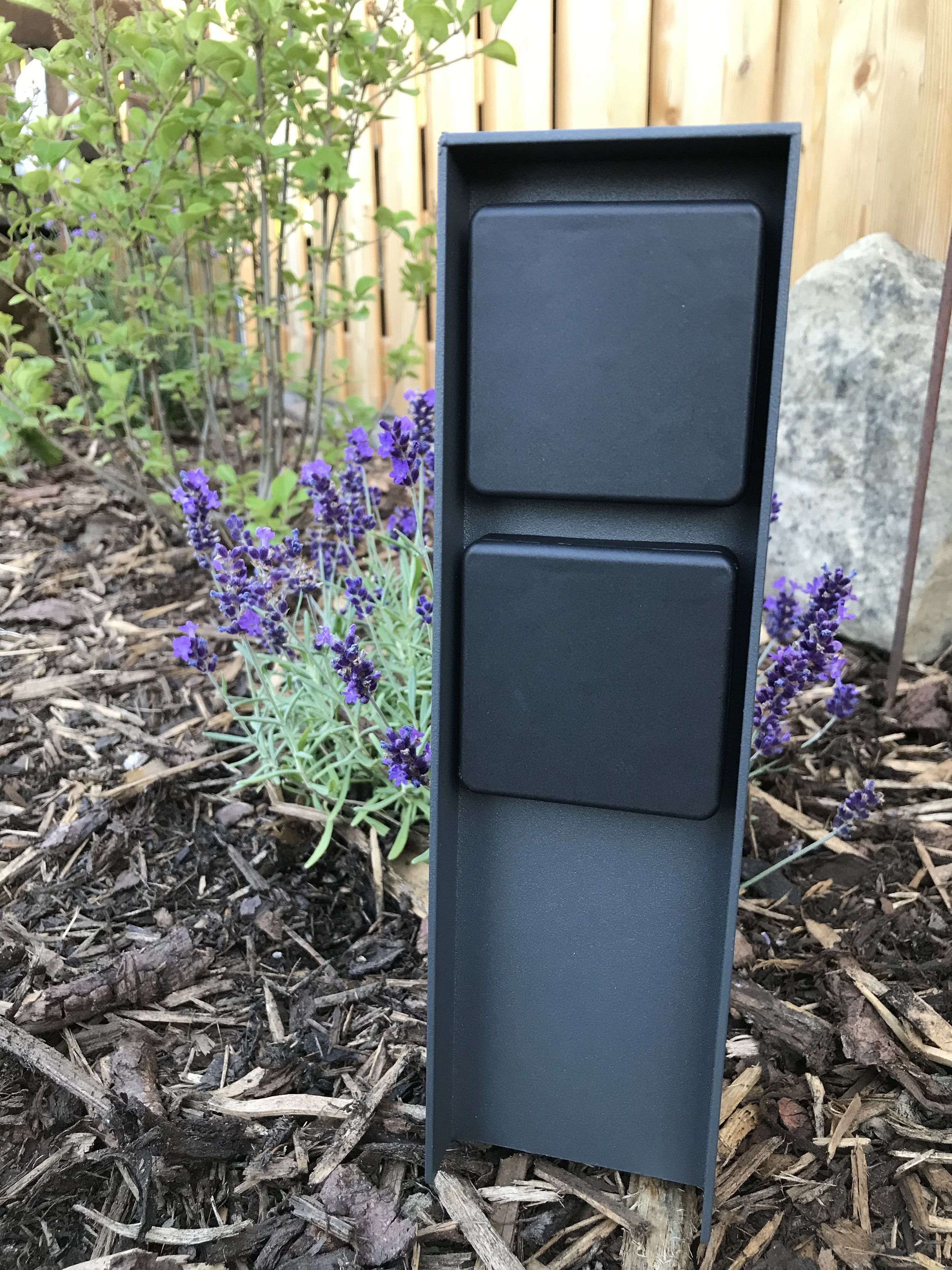 Schwarzer Außenpfosten mit zwei quadratischen Paneelen, gesetzt in einen Garten.