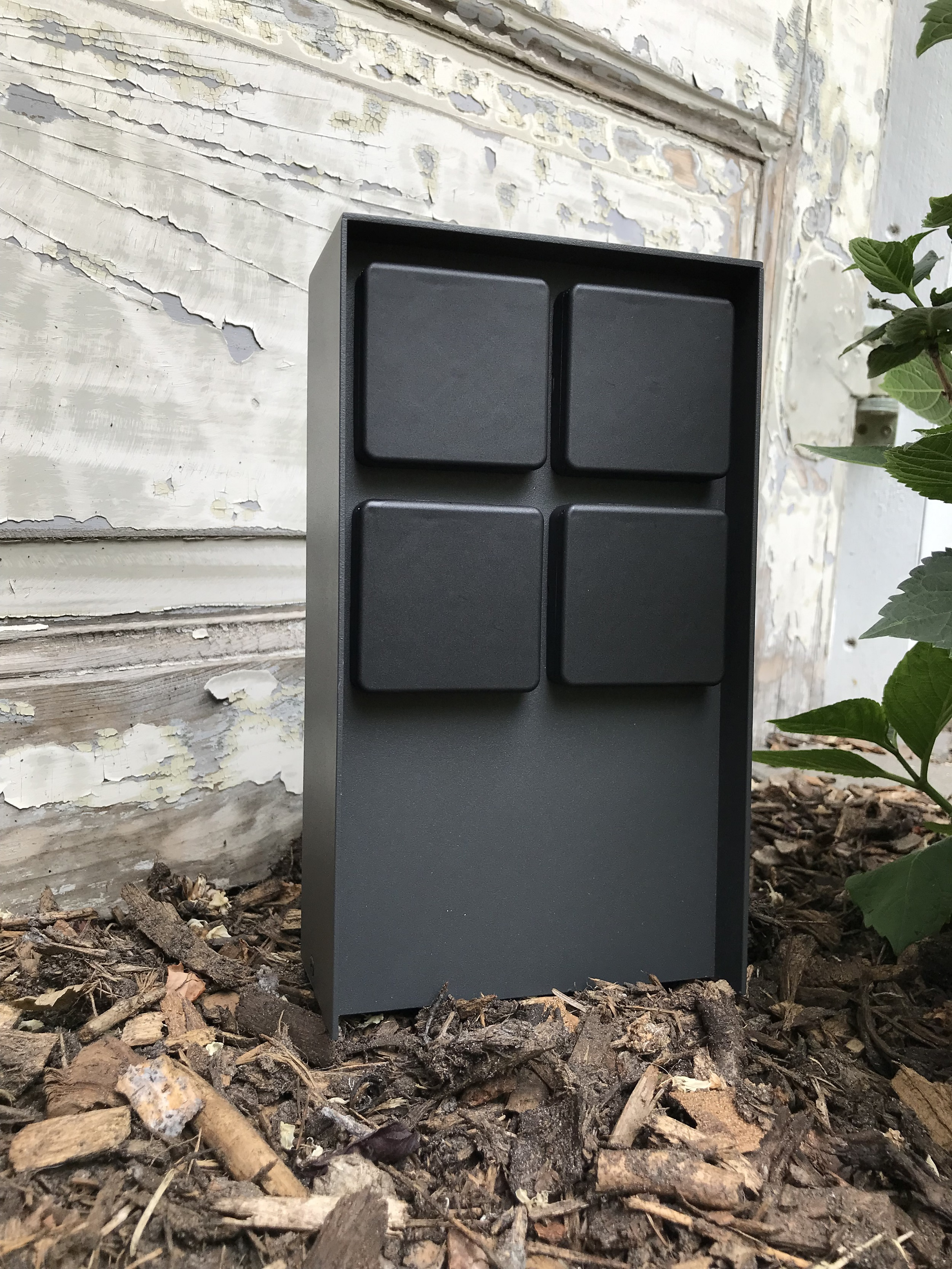 Dunkelgrauer Briefkasten mit vier quadratischen Öffnungen. Er steht auf einem Holzhackbett mit Pflanzen im Hintergrund.