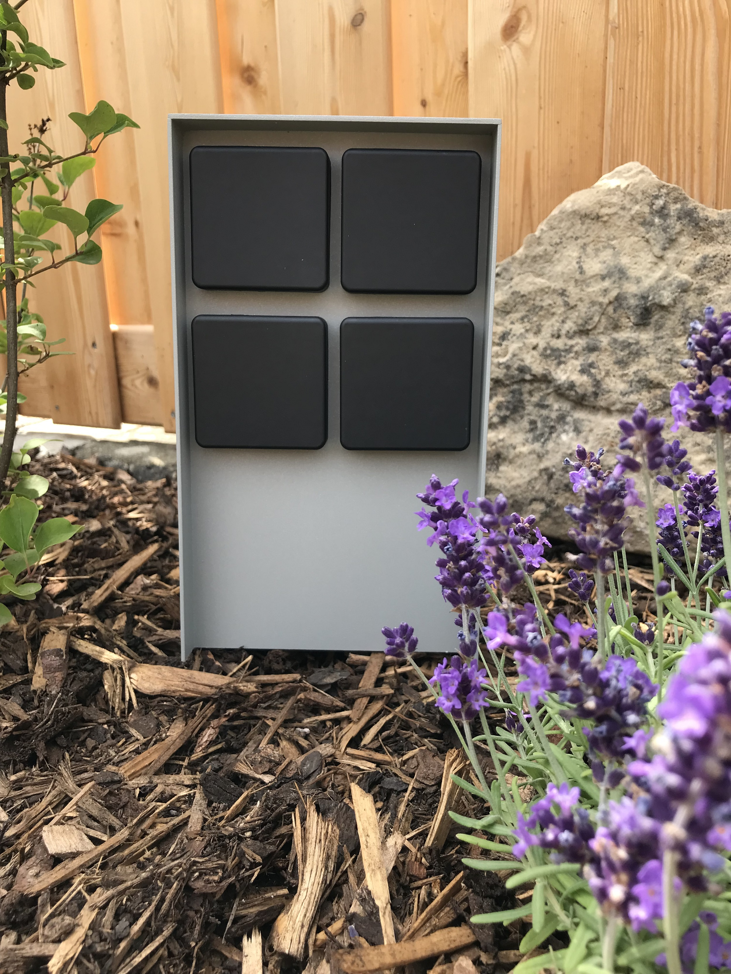 Grauer Metallkasten mit vier schwarzen Quadraten, in einem Garten mit lila Blumen und Holzspänen.