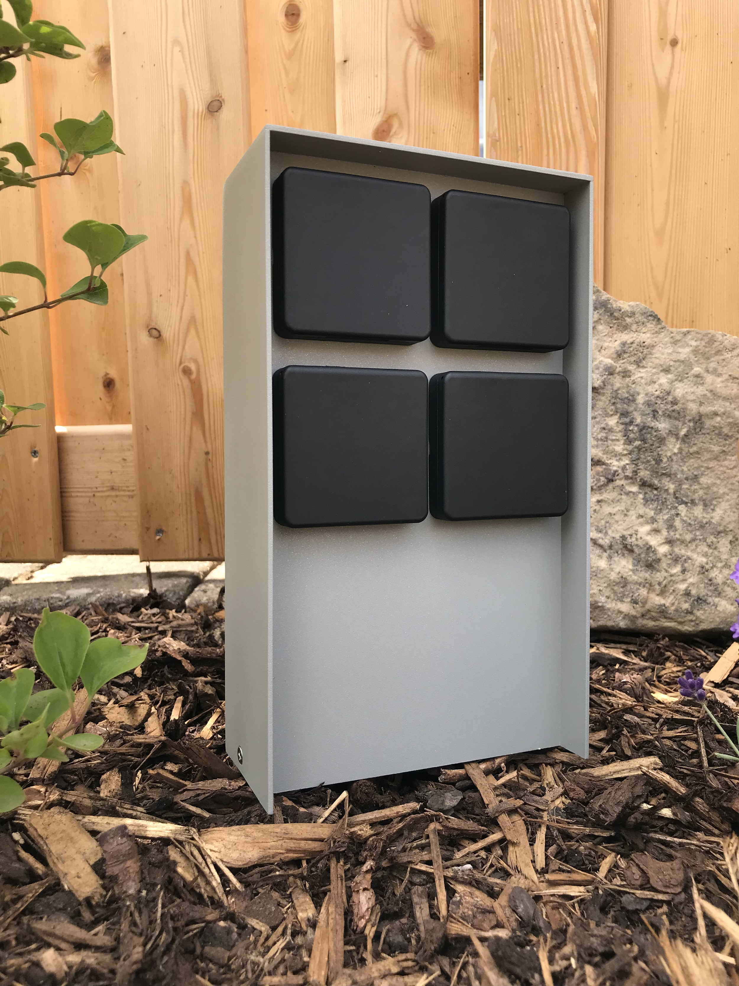 Eine graue elektrische Außenbox mit vier schwarzen quadratischen Abdeckungen. Die Box befindet sich in einem Garten.
