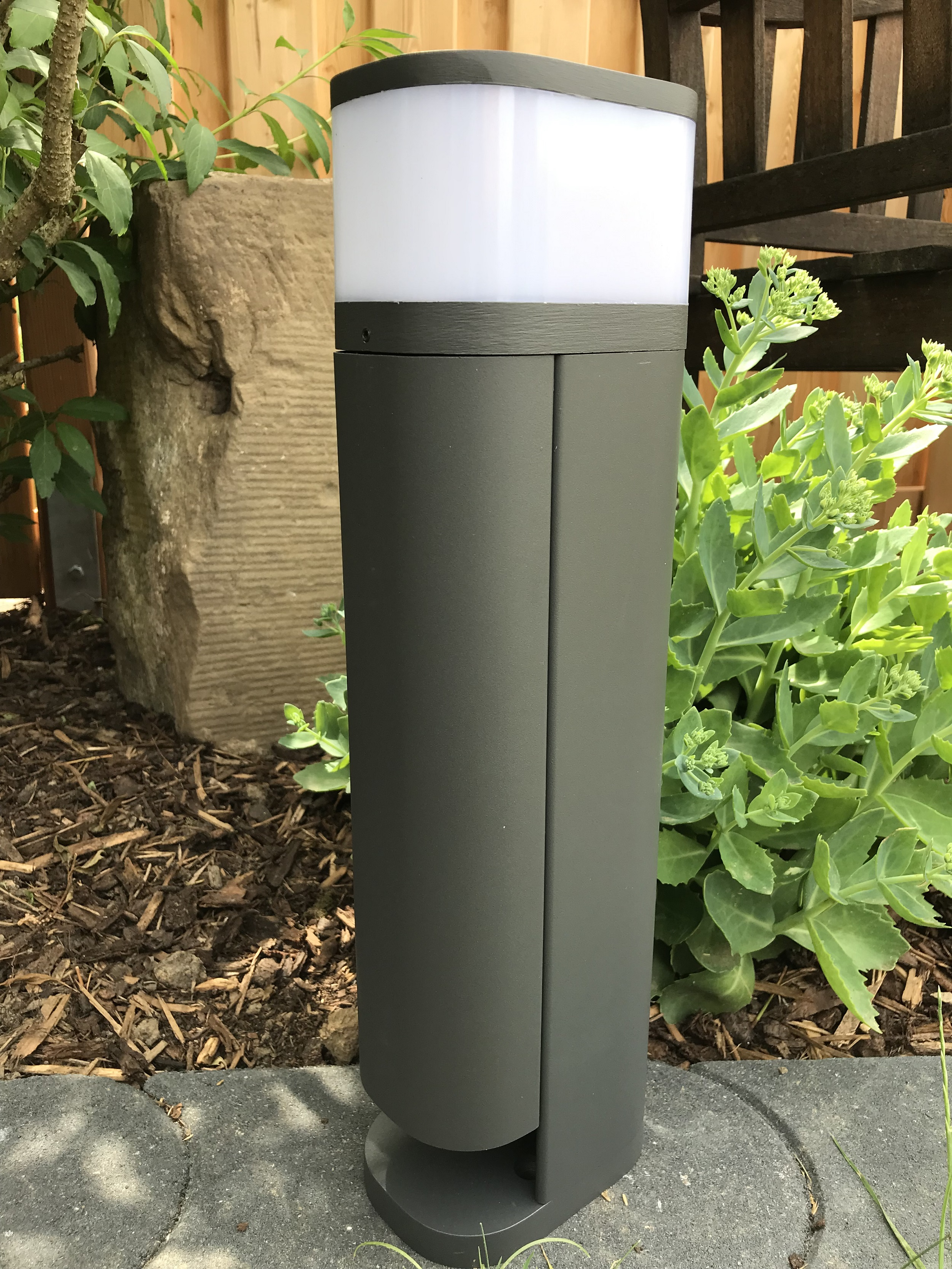 Eine graue Außenleuchte in einer Gartenszene. Sie hat einen weißen Kopf und einen dunkelgrauen Sockel.