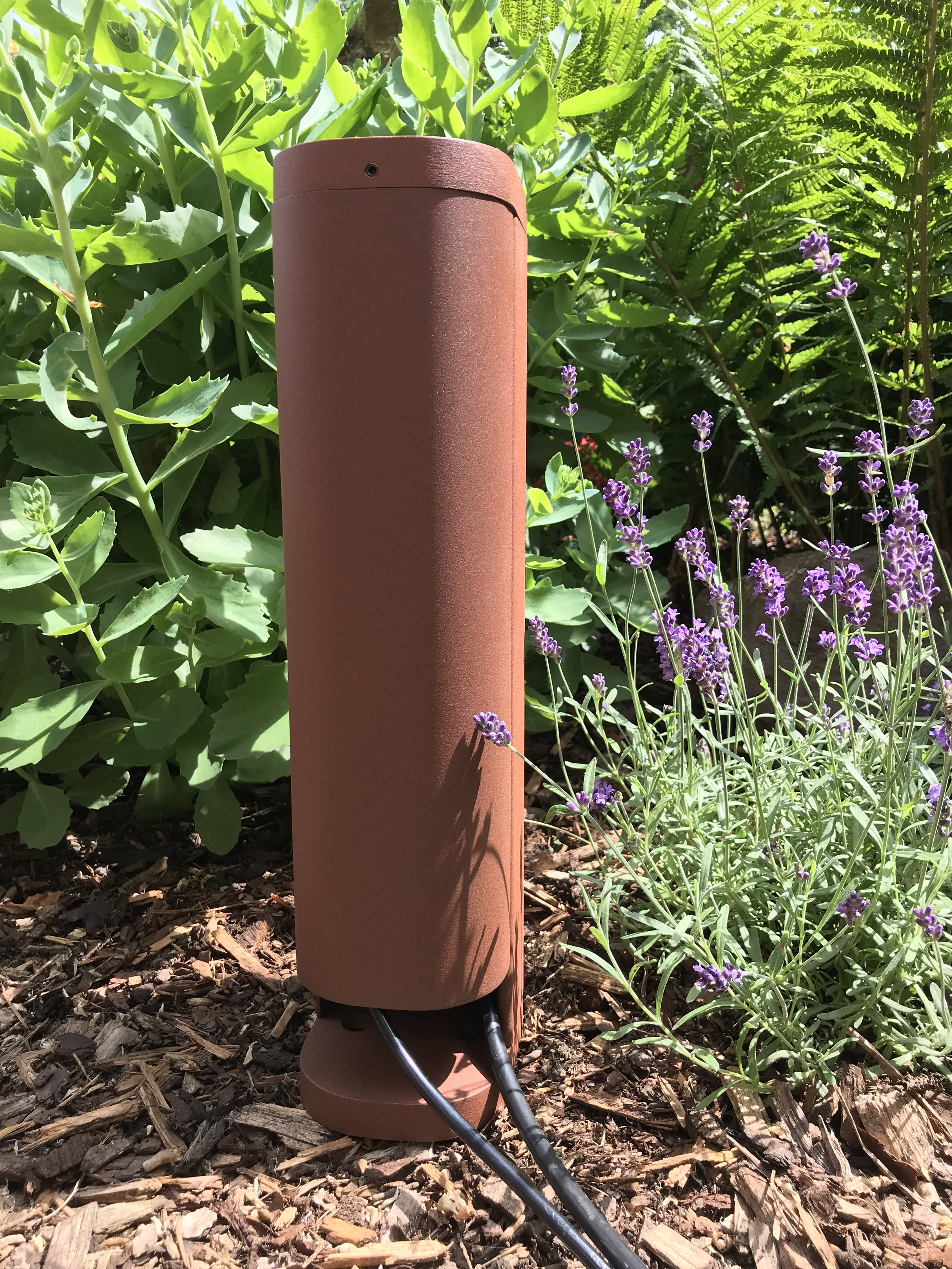 Un altoparlante cilindrico marrone in un giardino, con piante e fiori intorno.