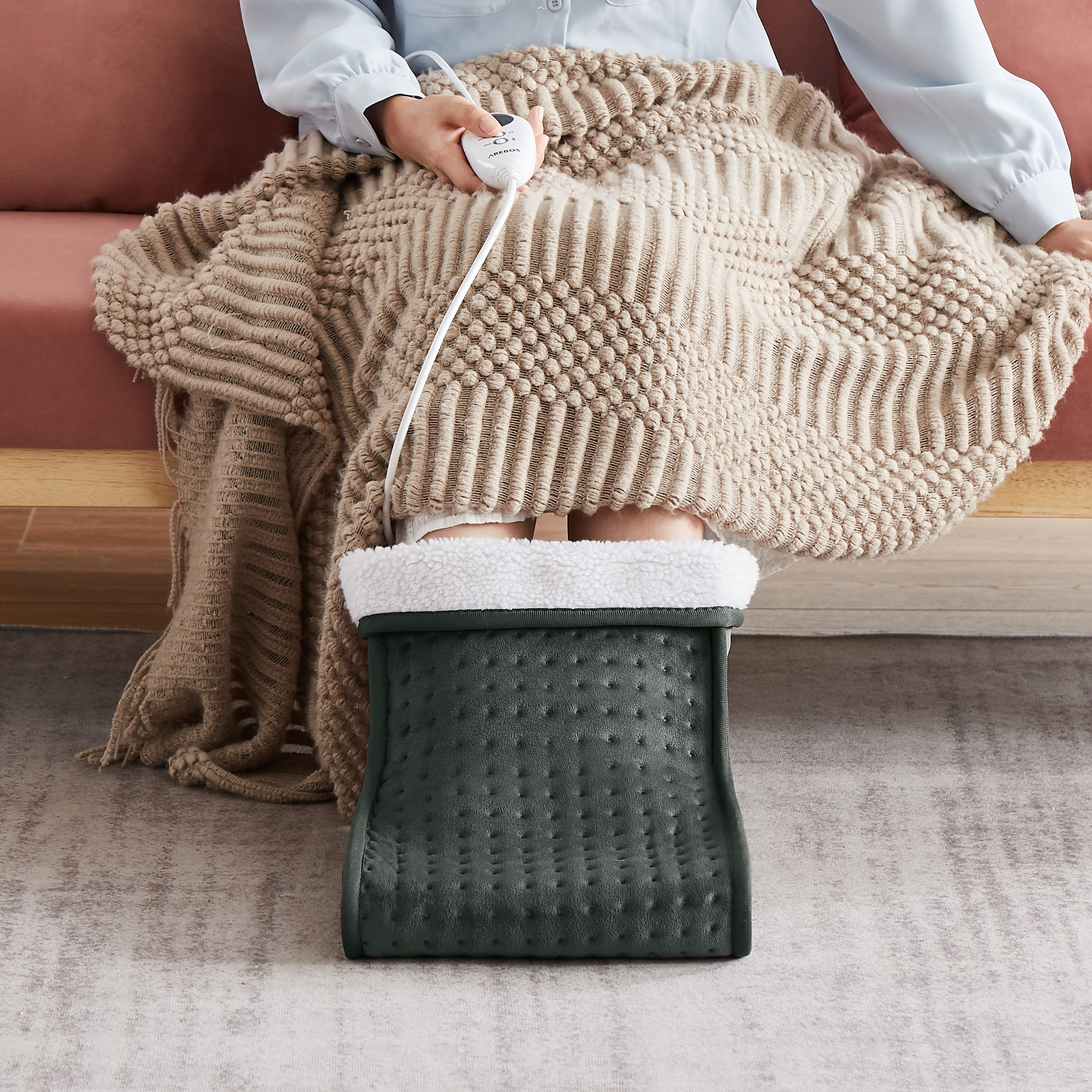 Person nutzt Fußwärmer, Decke über Beinen und Füßen, sitzt auf Sofa.