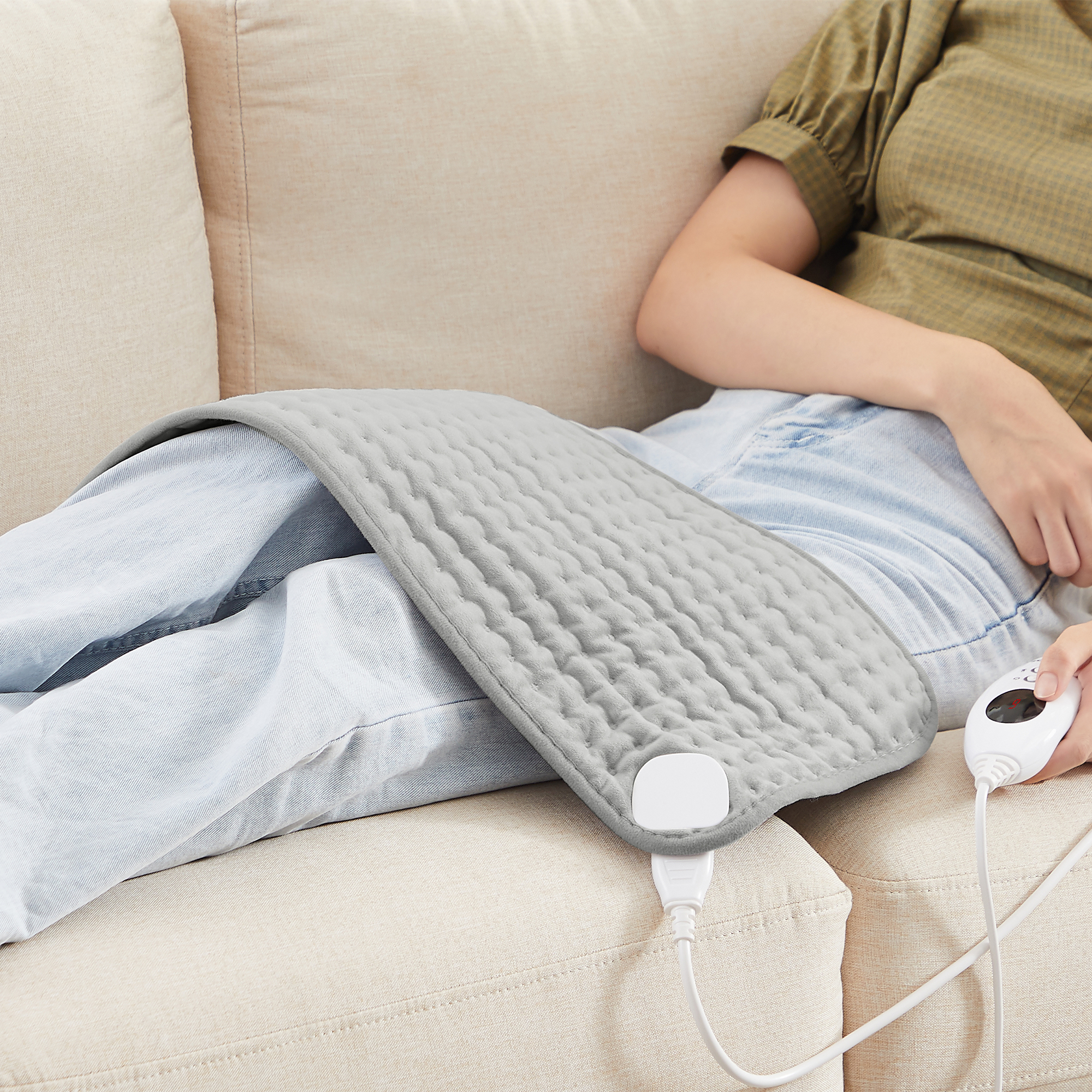 Eine Person liegt auf einer Couch mit einem grauen Heizkissen auf dem Bein und einer Fernbedienung.