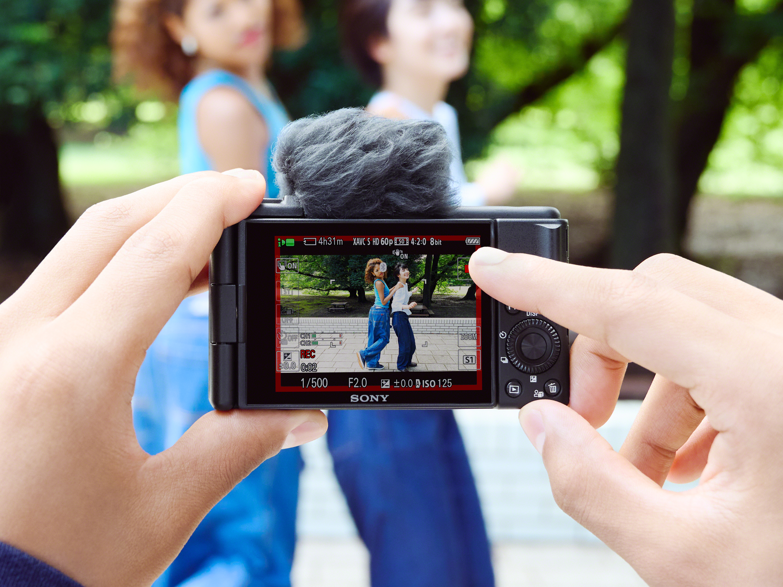 A person holding a camera, filming two people. Green trees are in the background.