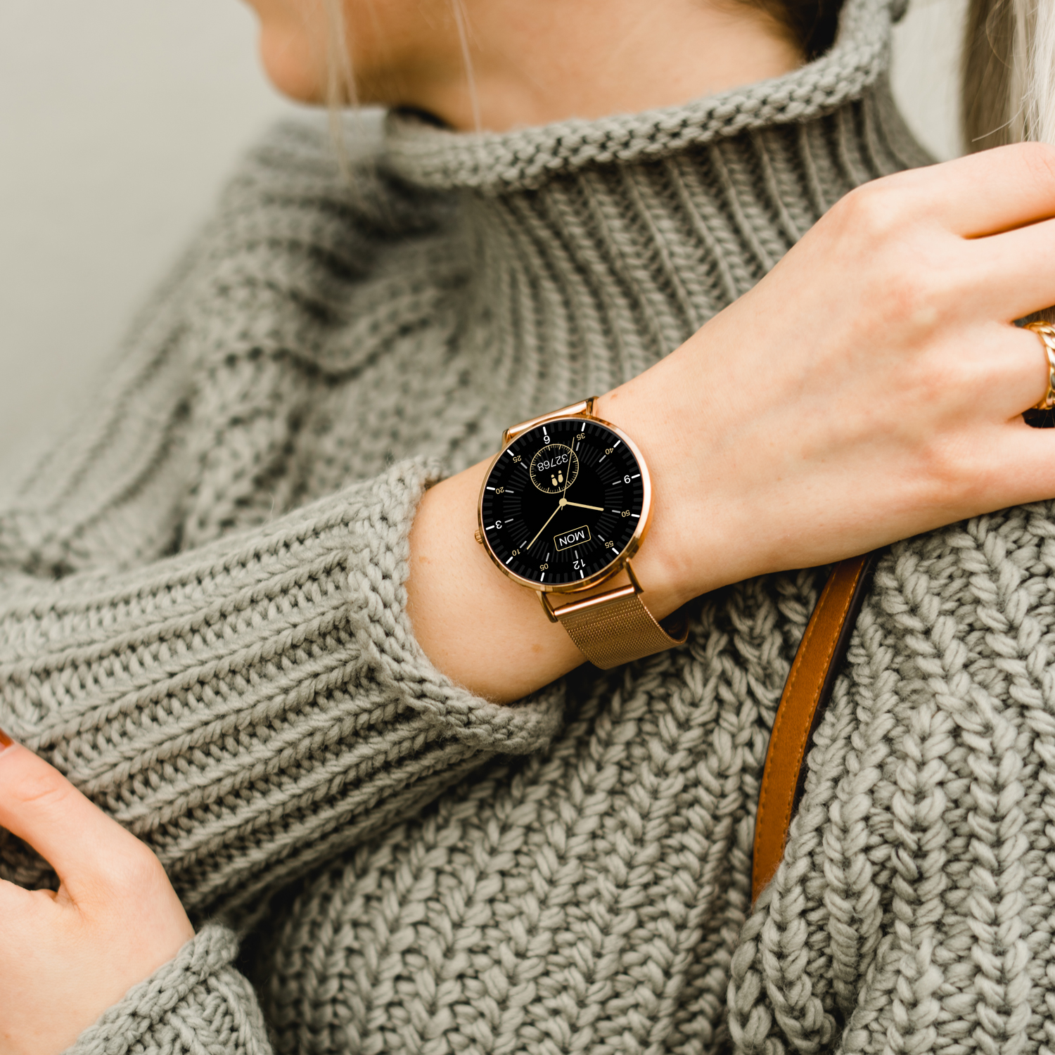 Frau mit grauem Pullover, zeigt goldene Uhr mit schwarzem Zifferblatt und goldenem Armband. Ring sichtbar.