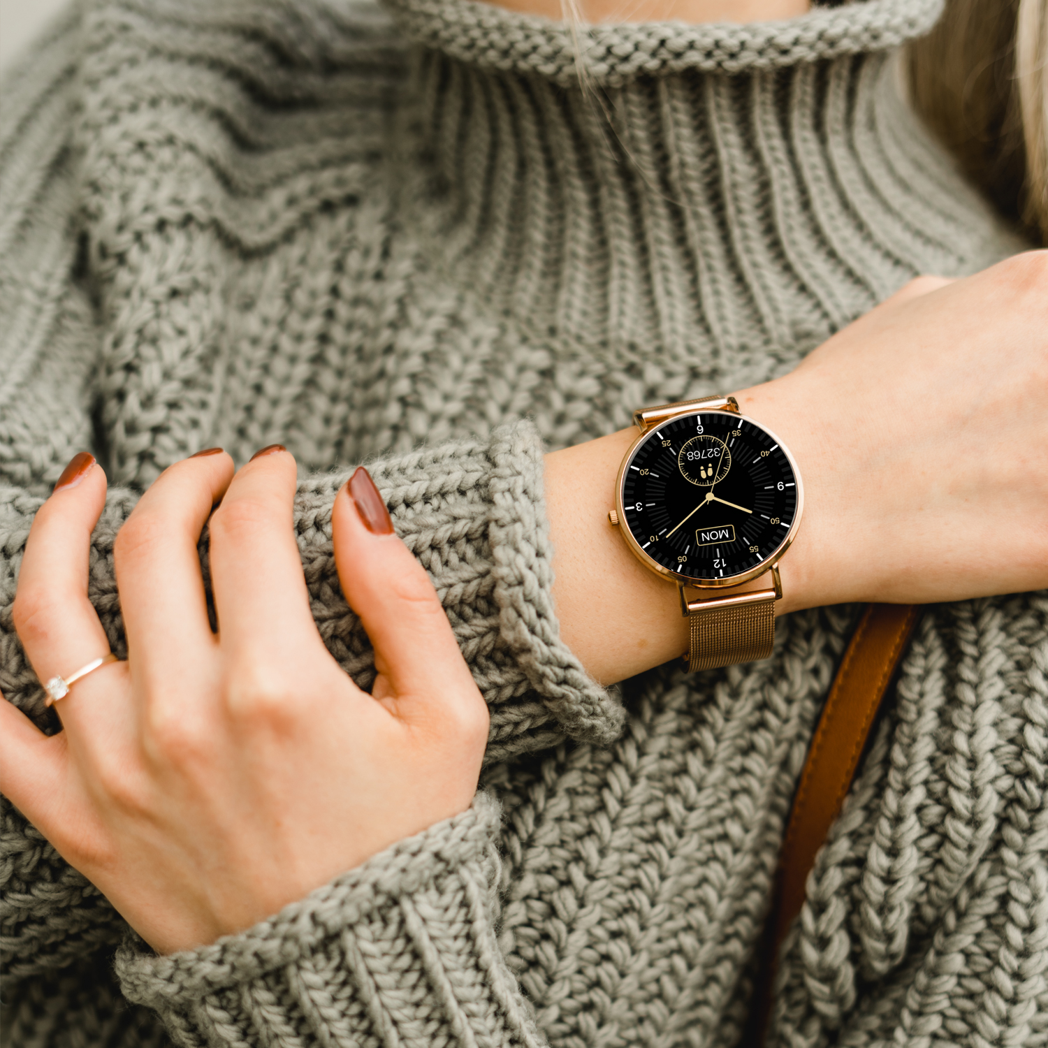 Handgelenk einer Frau mit goldener Uhr und Ring, trägt grauen Pullover.