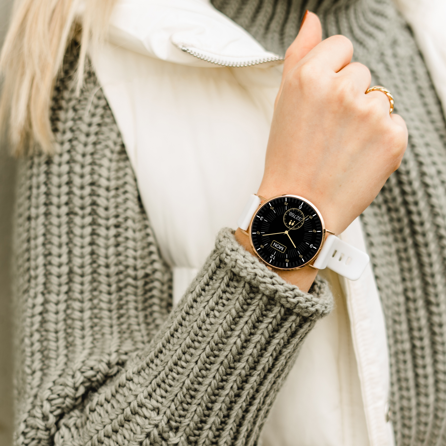 Handgelenk einer Frau mit goldener Uhr und weißem Armband. Sie trägt einen grauen Pullover und Weste.
