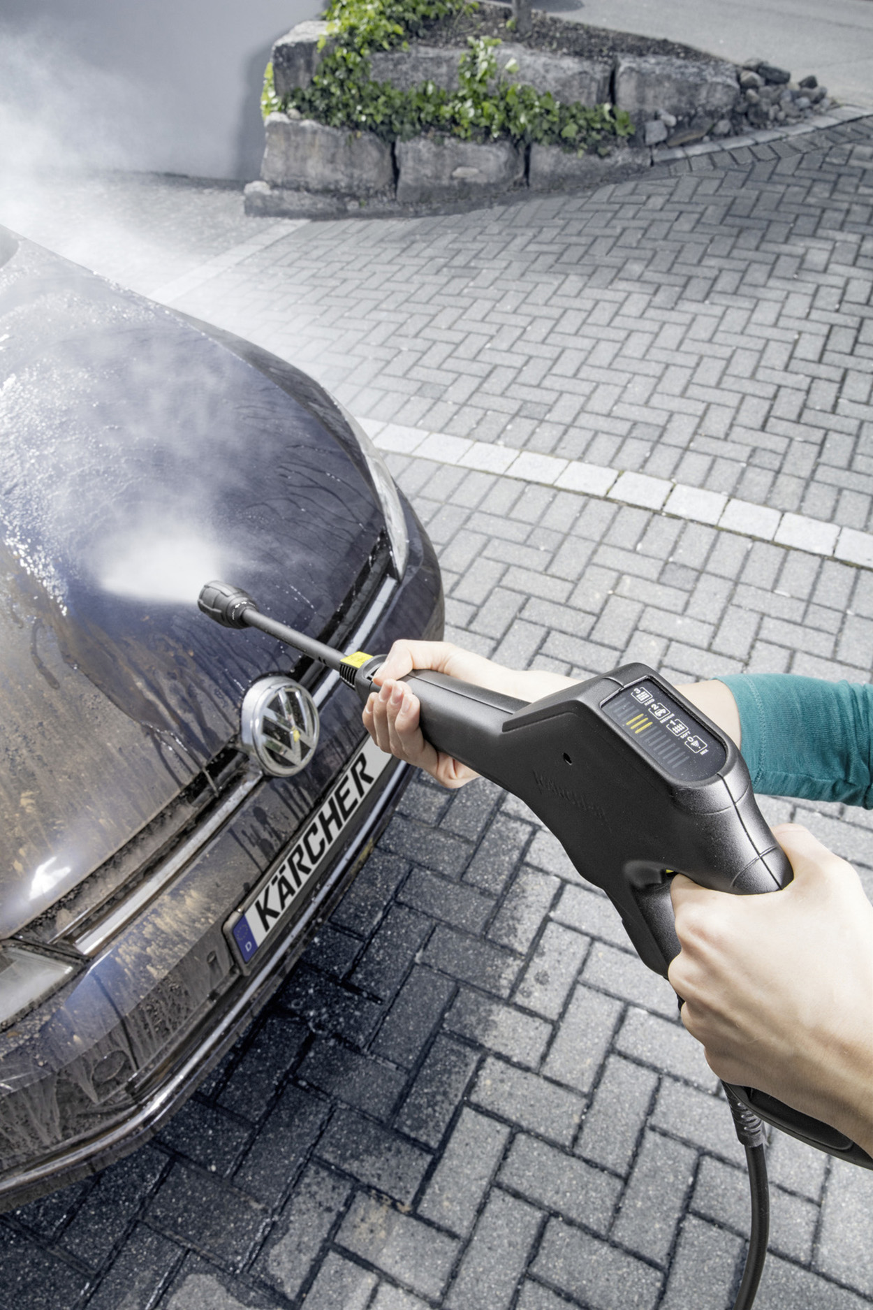 Person wäscht ein Auto. Ein Hochdruckreiniger besprüht die Motorhaube eines dunkelblauen Autos.