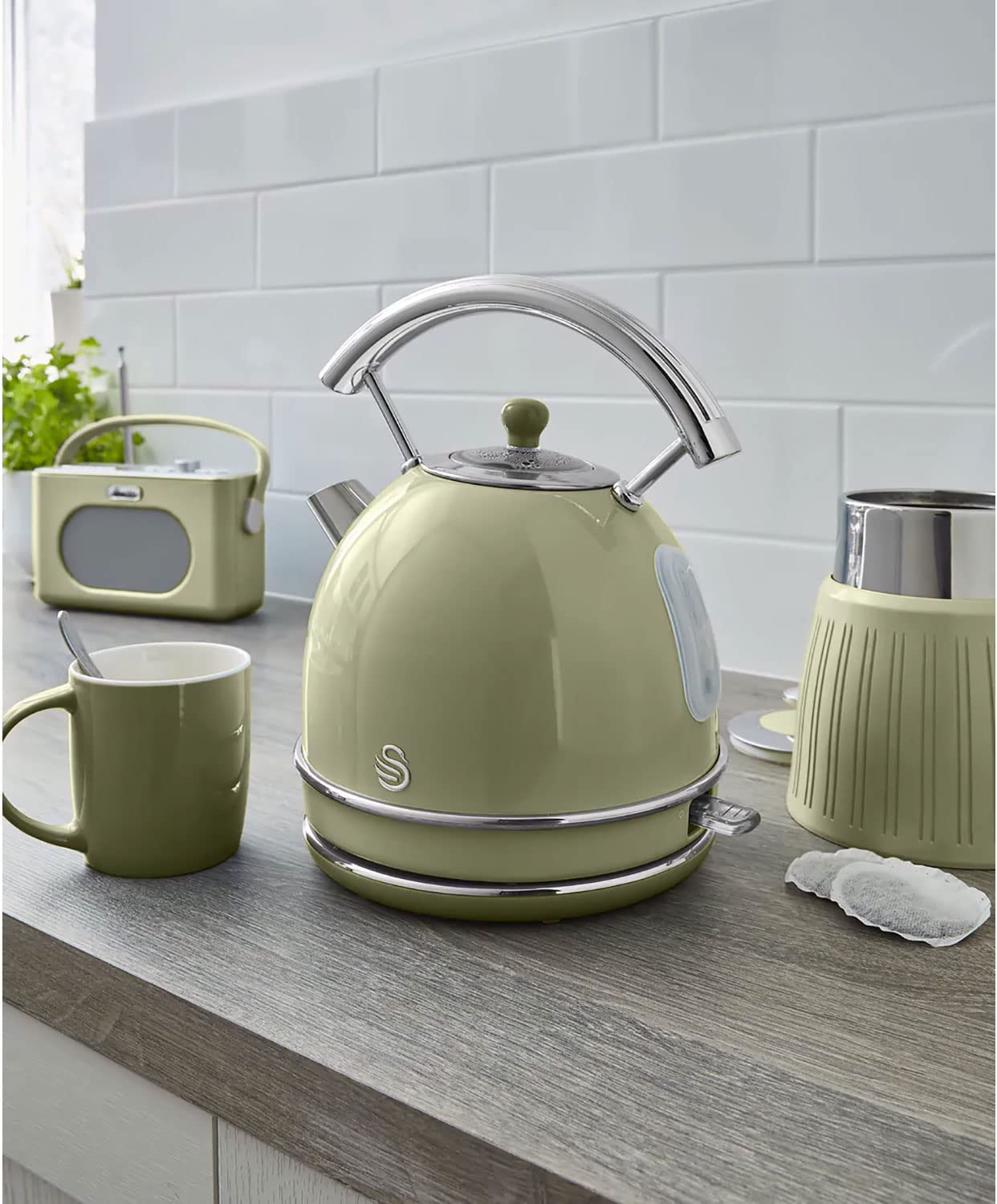 Escena de cocina con una tetera verde salvia, taza y otros elementos sobre una superficie de madera.