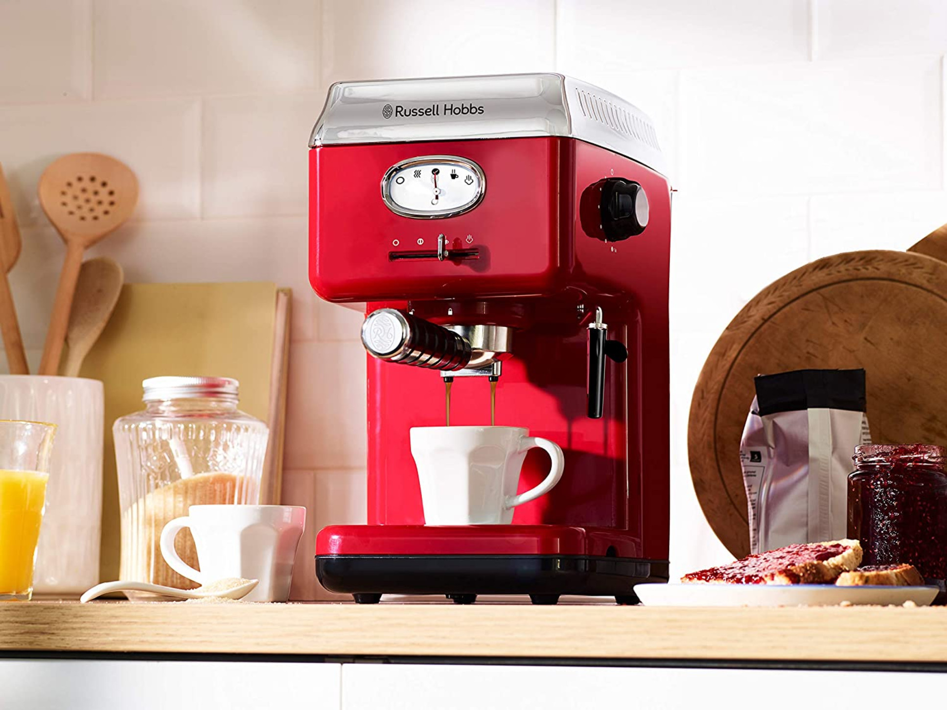 Eine rote Espressomaschine mit einer Tasse, die Kaffee auffängt. Andere Küchenartikel sind auf der weißen Arbeitsplatte sichtbar.